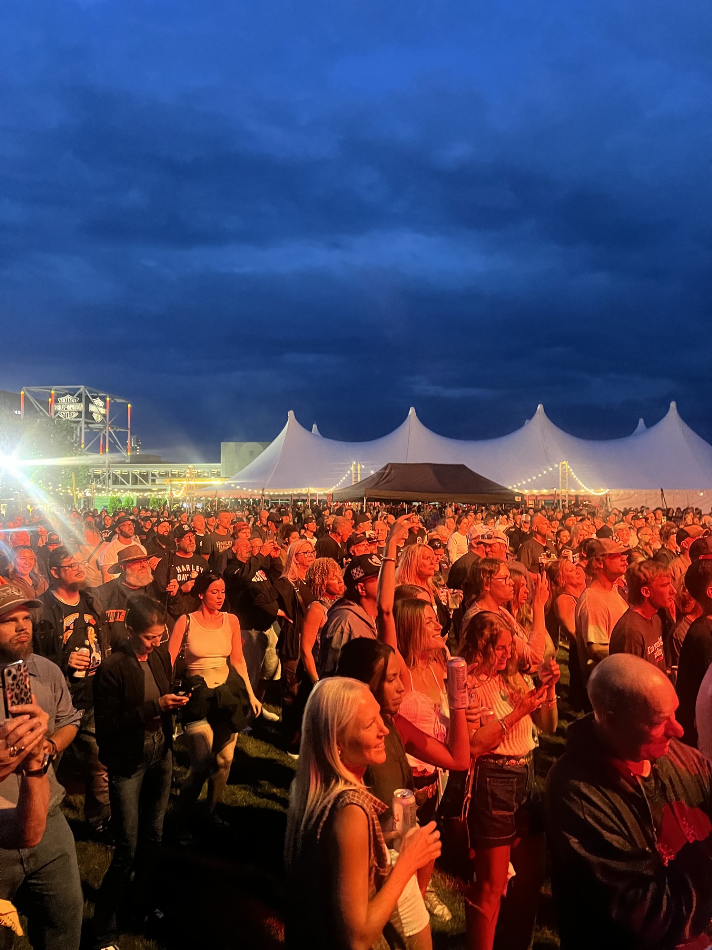 Gran multitud reunida en concierto al aire libre cerca de carpas blancas bajo el oscuro cielo de la tarde