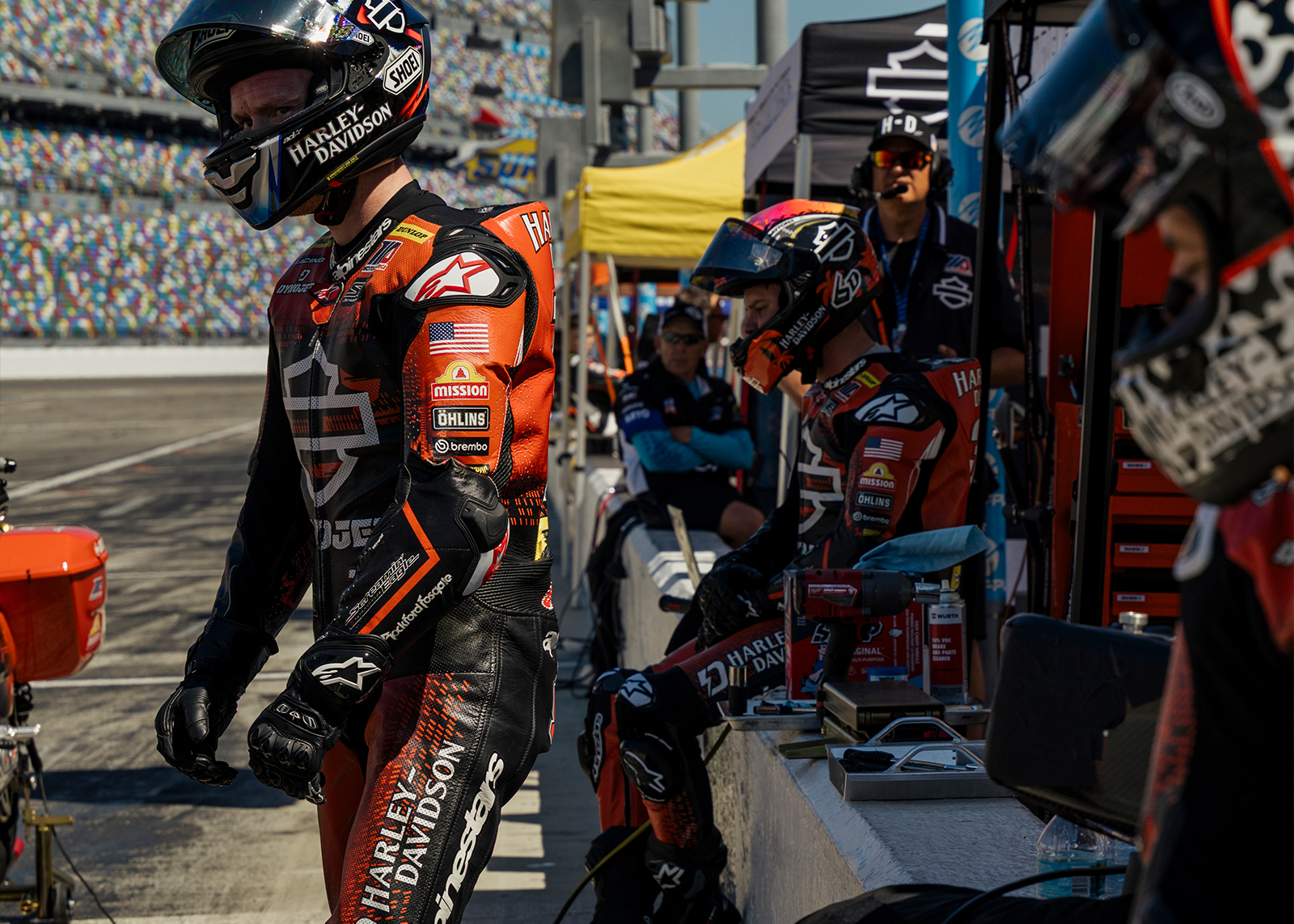 H-D factory racers stand or sit on the pit road wall waiting to mount their machines 