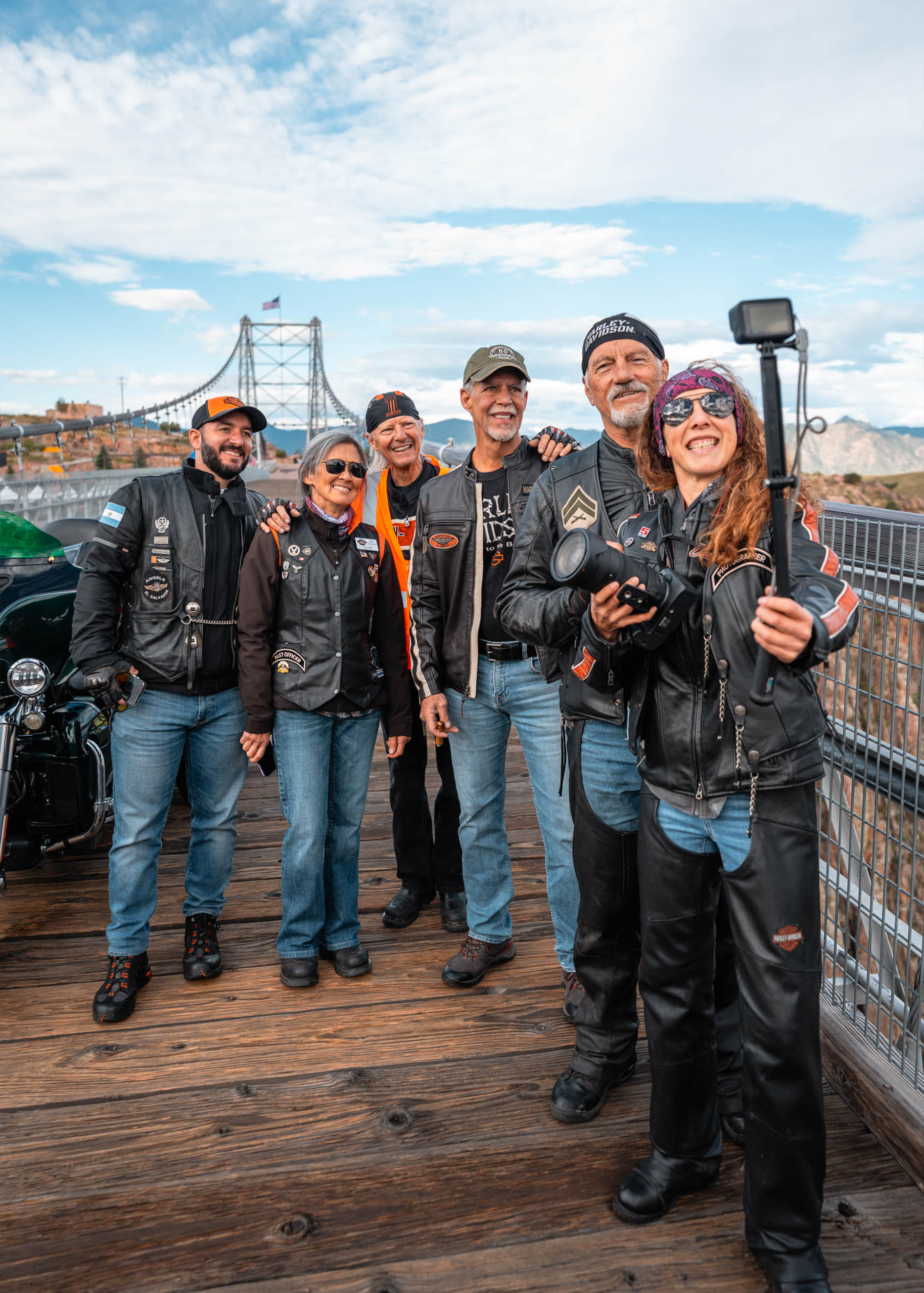 A group of H.O.G. members smile and pose on the Royal Gorge bridge as the person in front holds a selfie stick up to capture a photo.