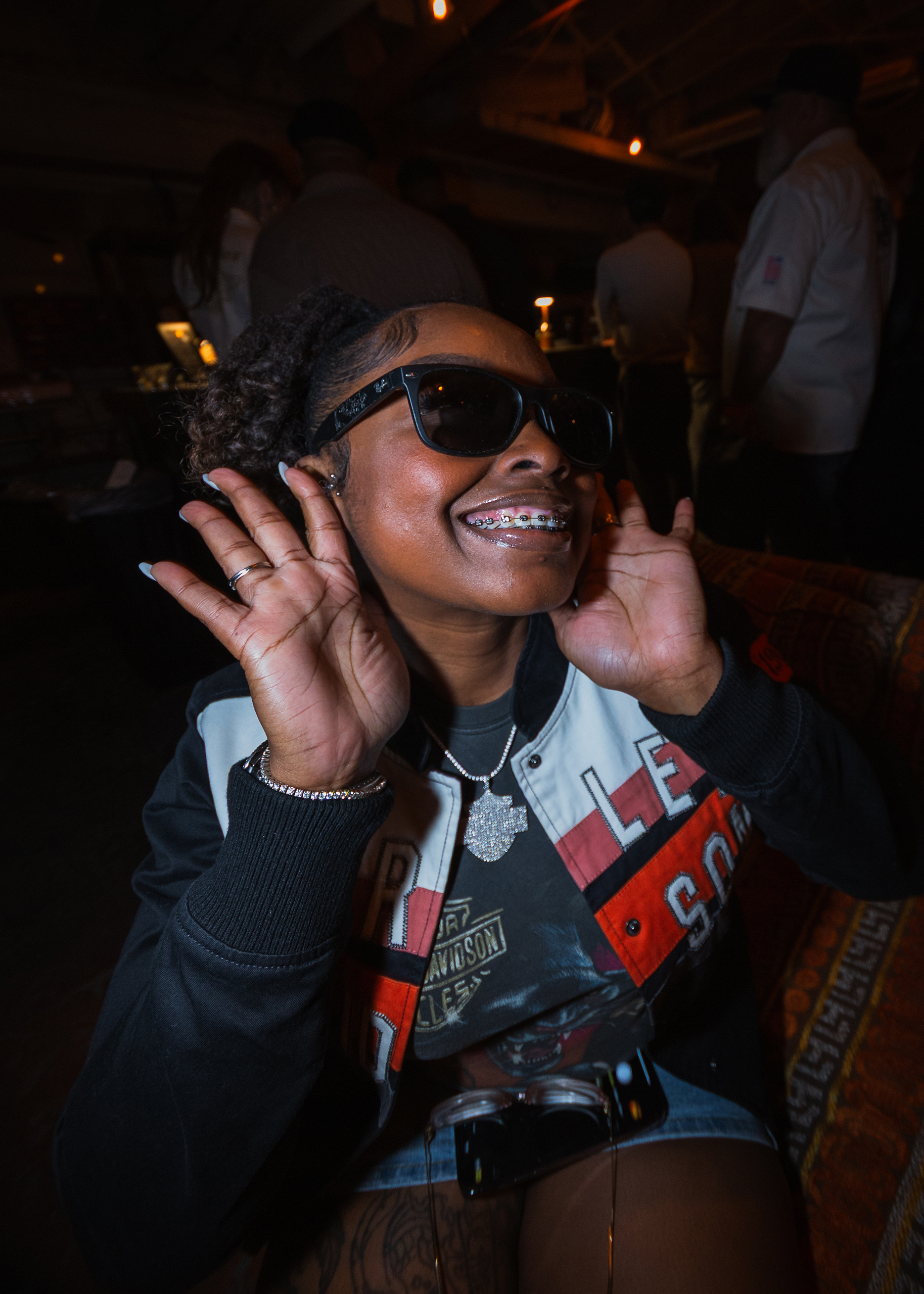 A woman in sunglasses, a Harley shirt, jacket, and bedazzled necklace uses her hands to frame her smiling face.