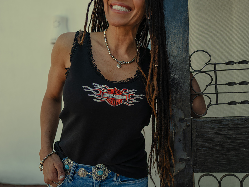 Woman smiling in lace-trimmed black tank top with bar & shield logo and flames