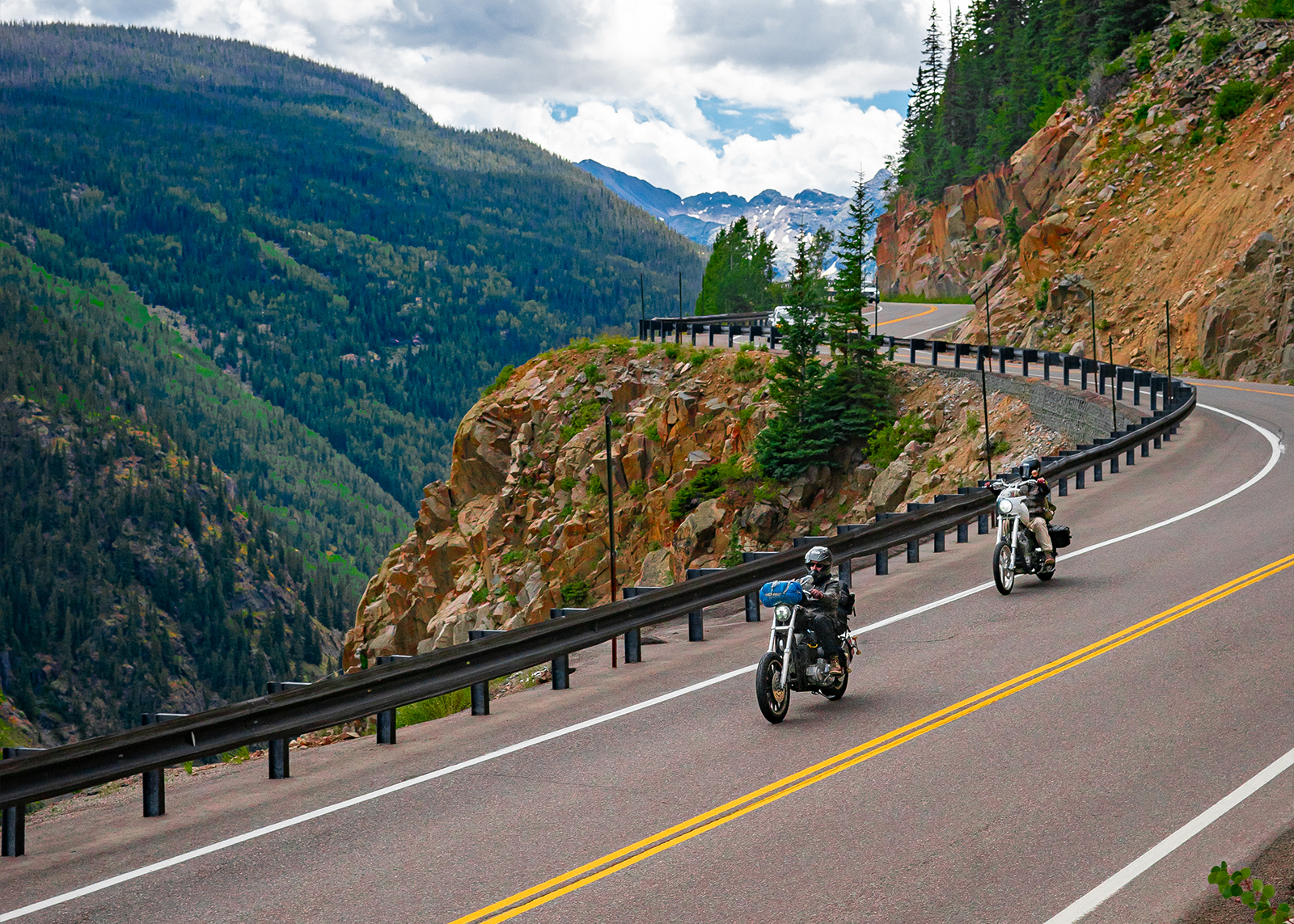 Two people ride vintage choppers on a cliffside highway in the mountains 