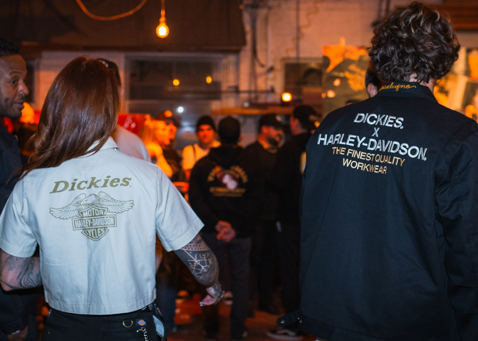 A man and woman, wearing a black HD x Dickies jacket and white HD x Dickies shirt respectively, shot from behind looking into a crowd of people. 