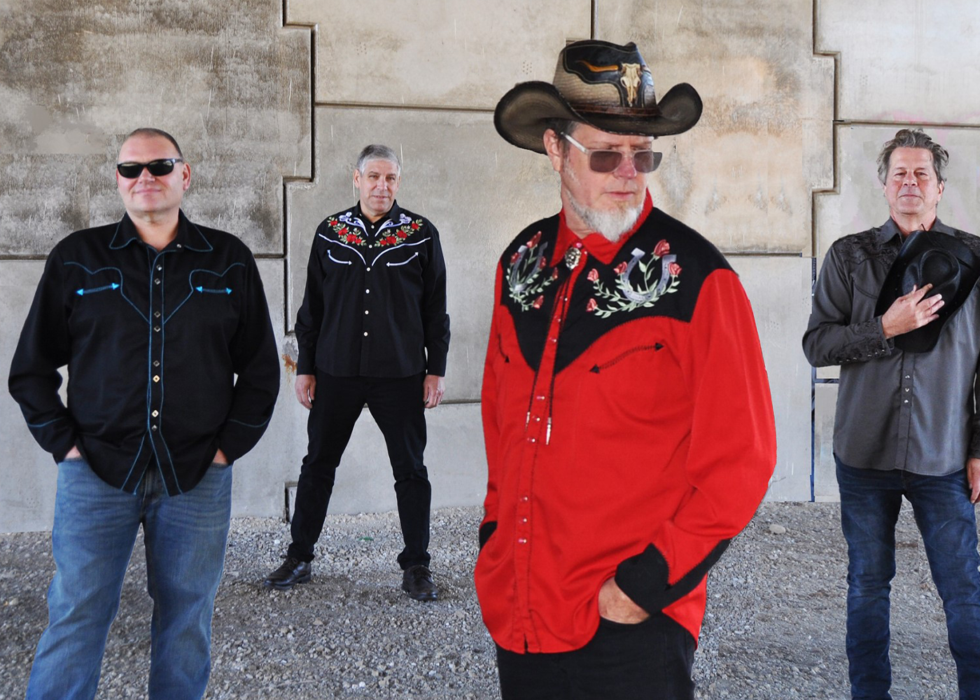Four band members standing against a concrete wall, one in red shirt and cowboy hat