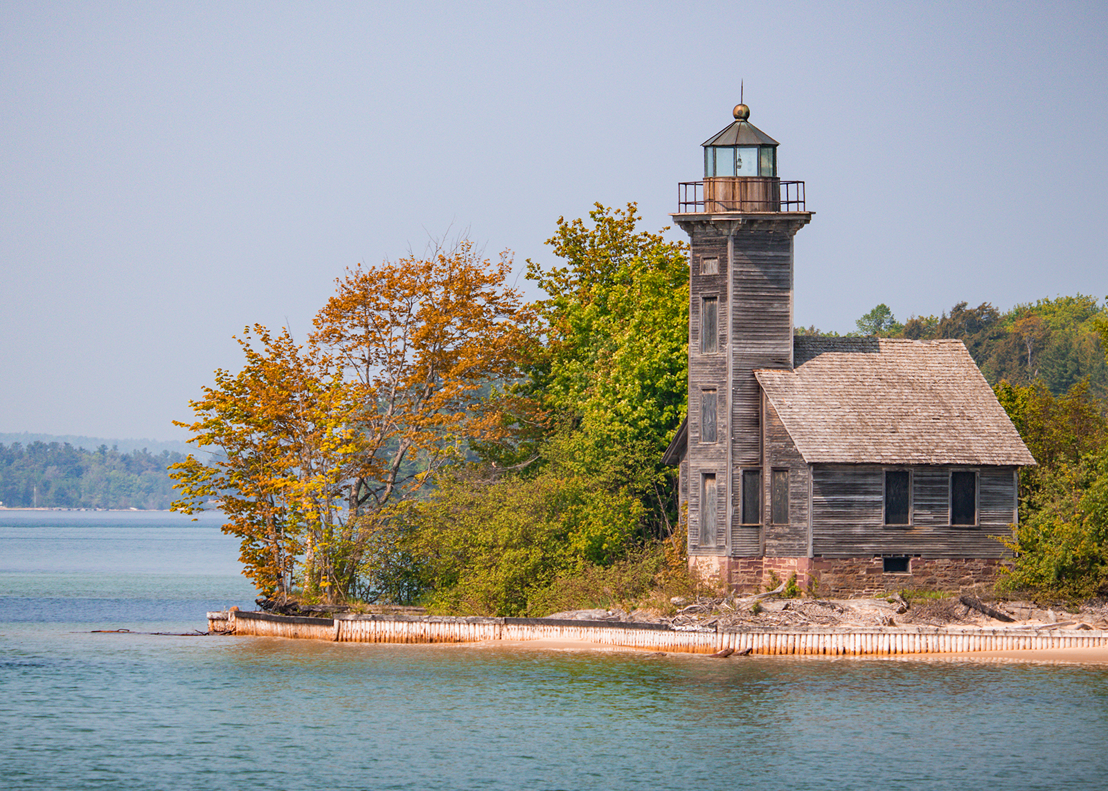 Old wooden lighthouse with attached house stands by the shoreline, surrounded by autumn-colored trees.