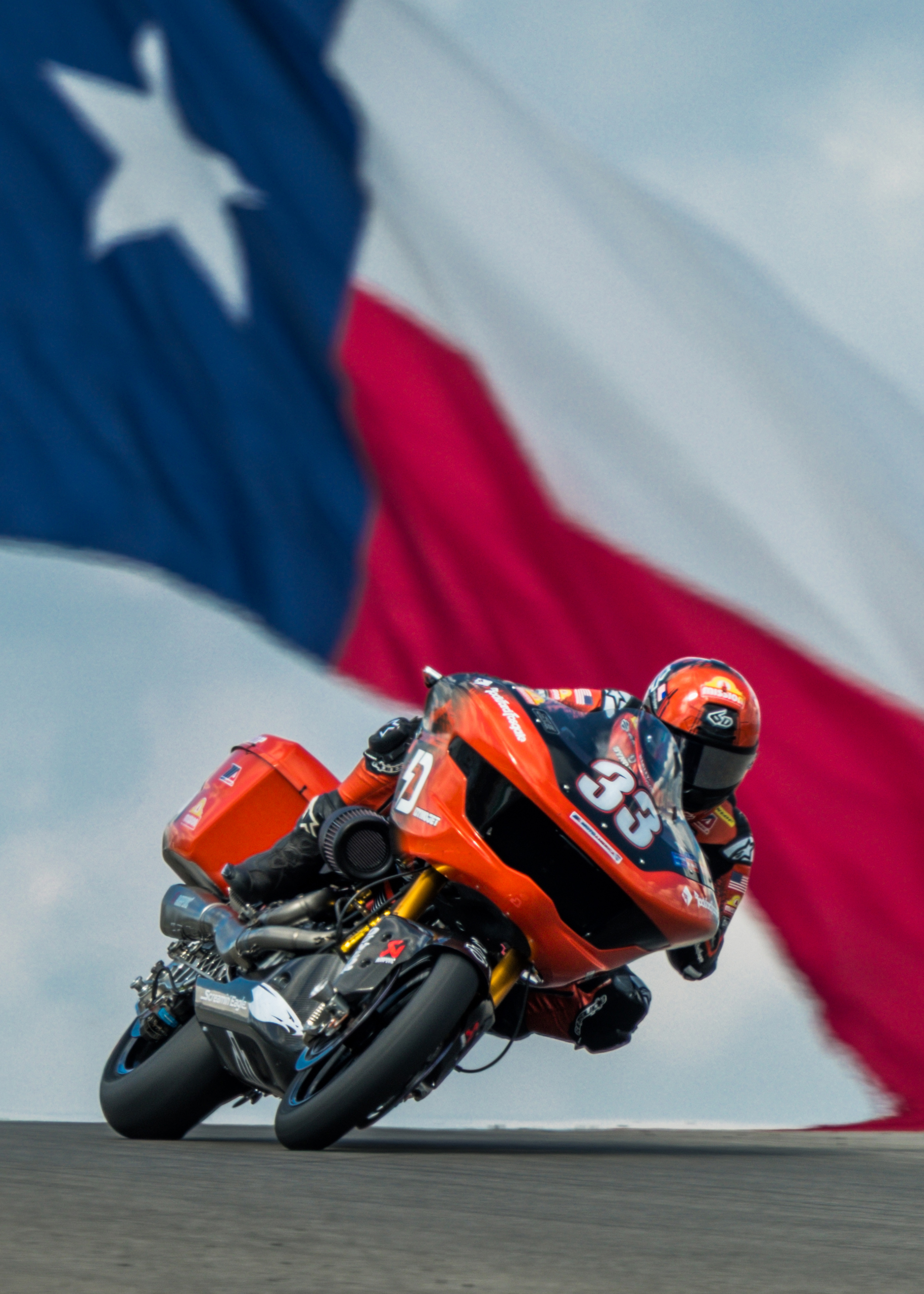 Kyle Wyman leans through a corner coming over a hill with a large Texas state flag waving in the background.