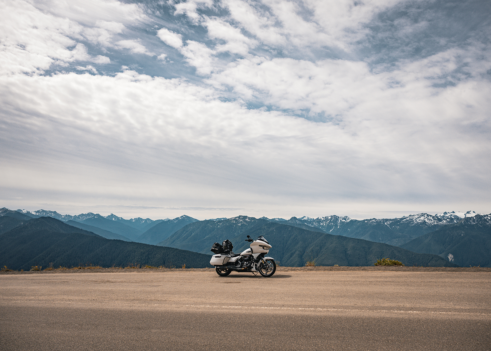 A white touring motorcycle parked on a mountain road with expansive snow-capped peaks in view.