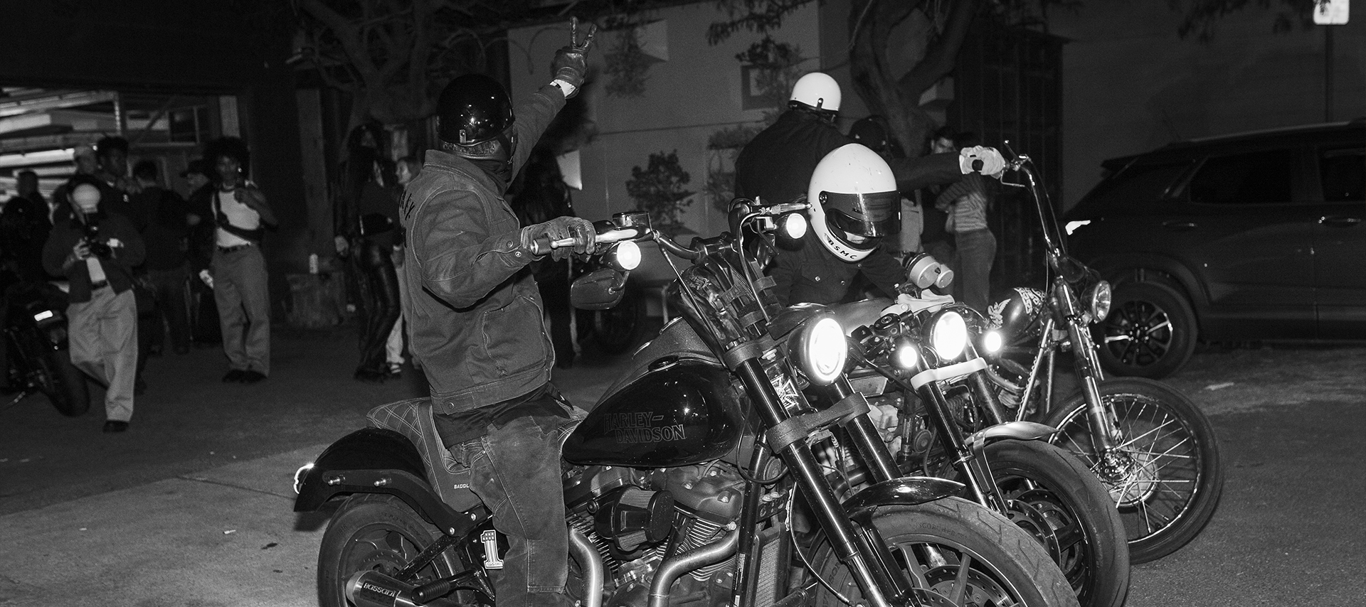 A person on a motorcycle turns around to give the peace sign to the crowd behind him, while two riders next to him prepare their bikes. 