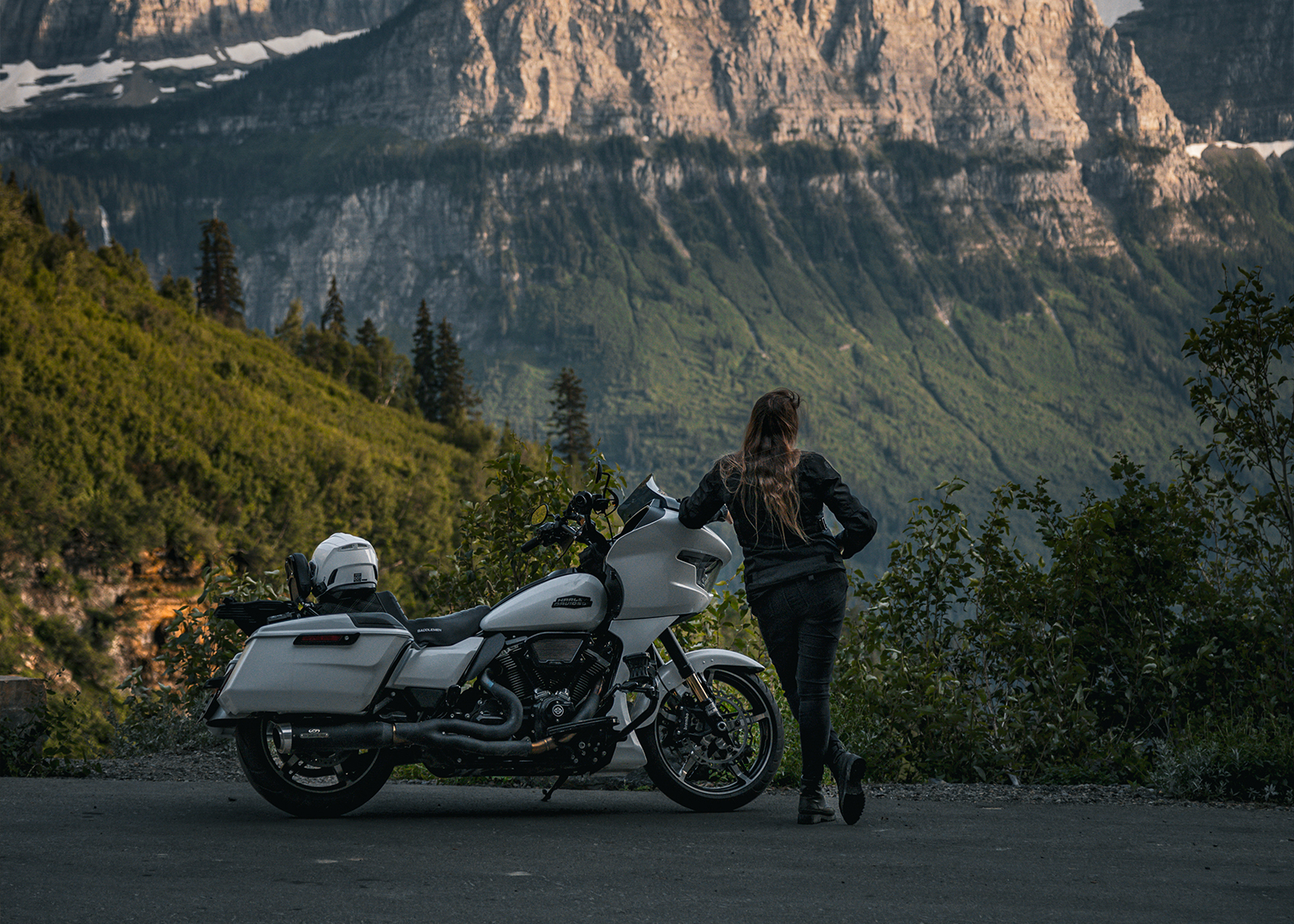 The author stands next to a white Harley-Davidson Road Glide motorcycle on a mountain roadside with her back to the camera.