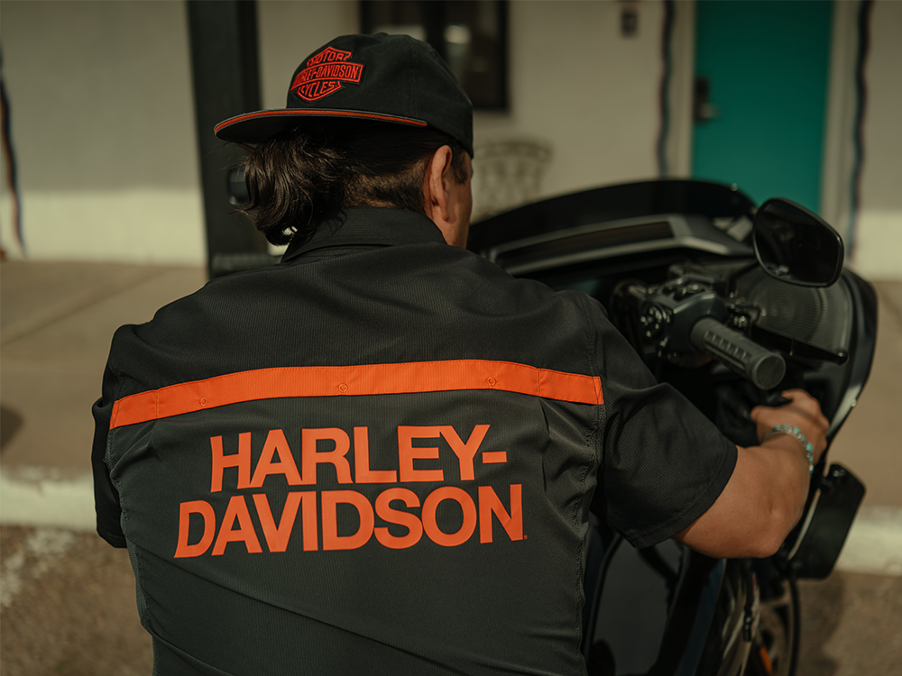 Man sitting on motorcycle with Harley-Davidson in orange across back of shirt and a bar & shield logo baseball hat