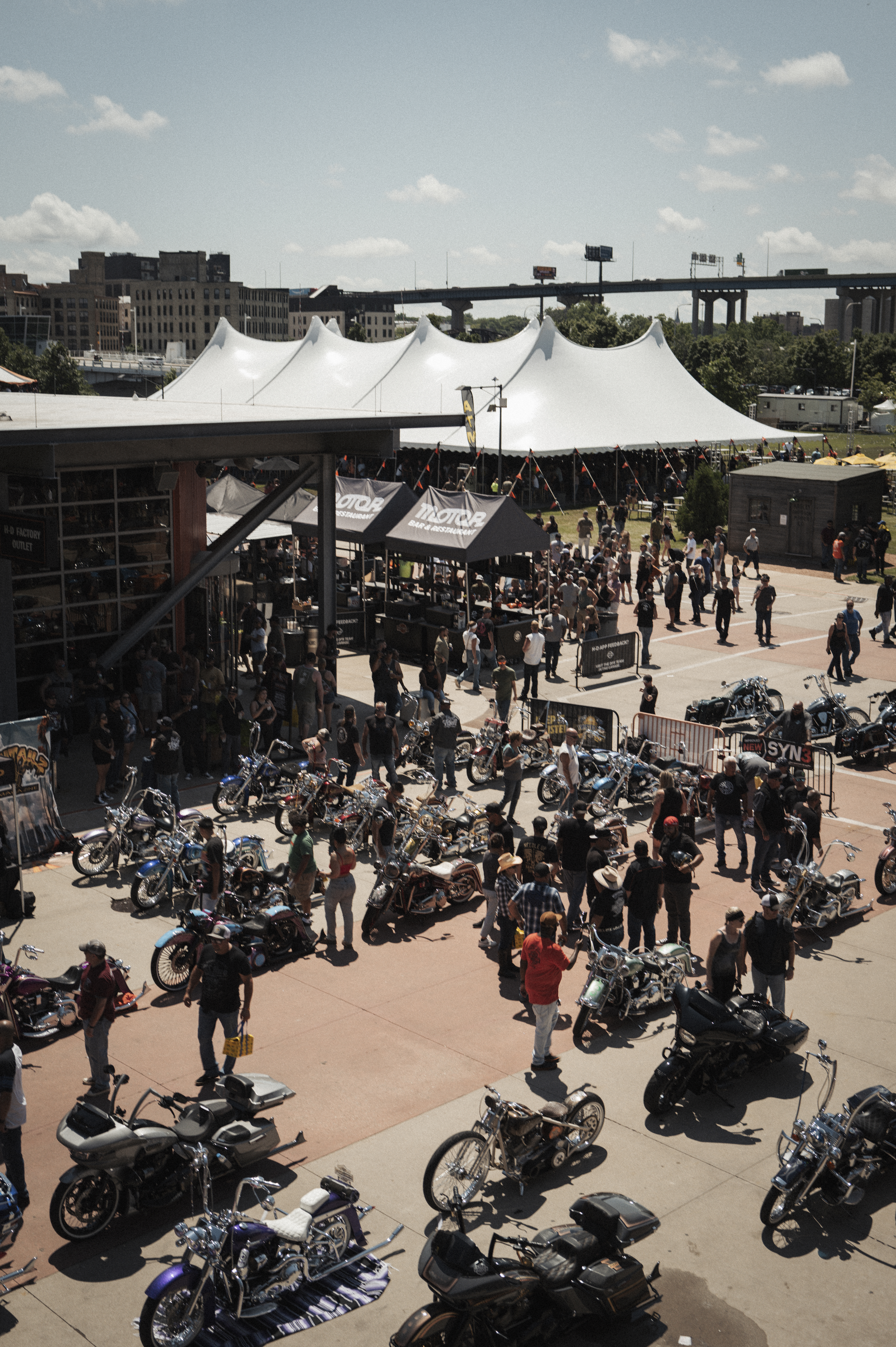 Plaza de eventos al aire libre concurrida con carpas, puestos de vendedores y motocicletas estacionadas.
