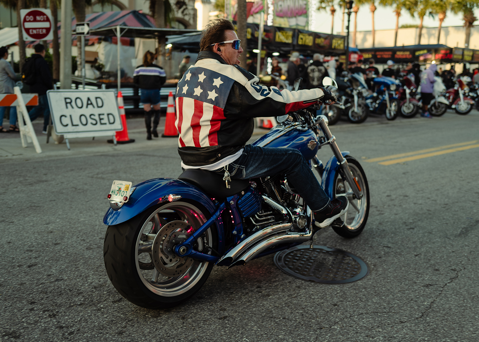 A man in an American Flag themed leather jacket rides his customized motorcycle down Main Street