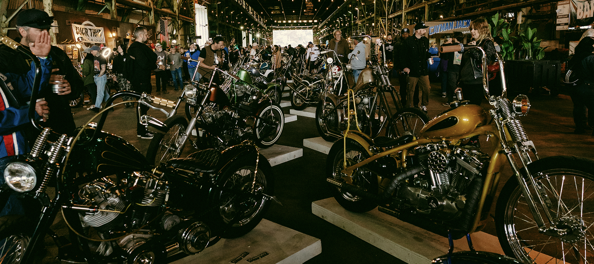 Two rows of bikes for viewing, one on the left and the other on right, lead down the middle of the building, with dozens of people walking on either side admiring the bikes. 