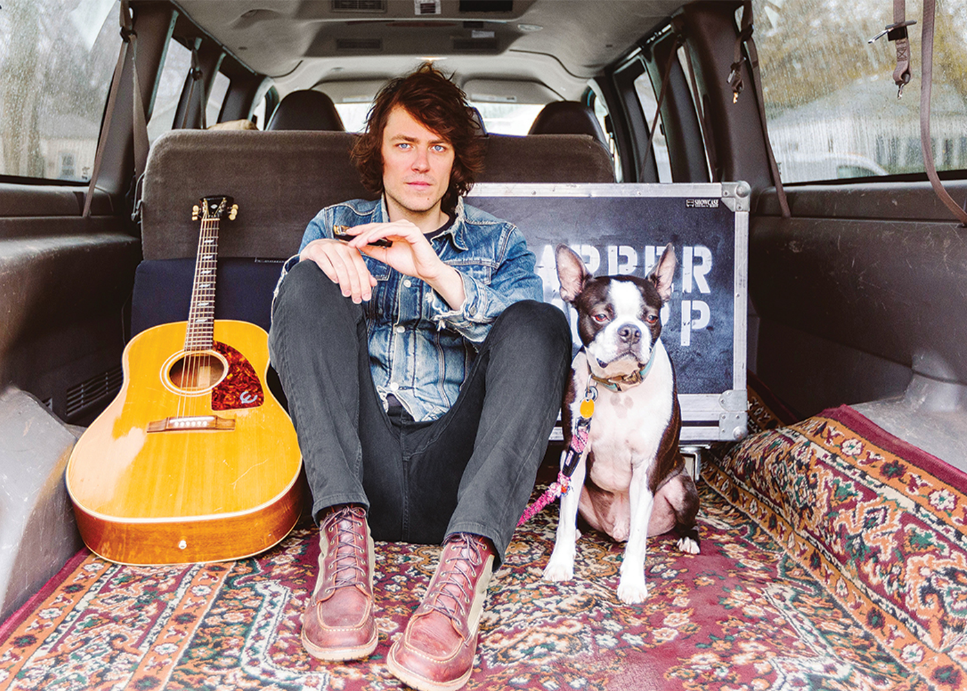 Person sitting in van with acoustic guitar and small dog on patterned rug