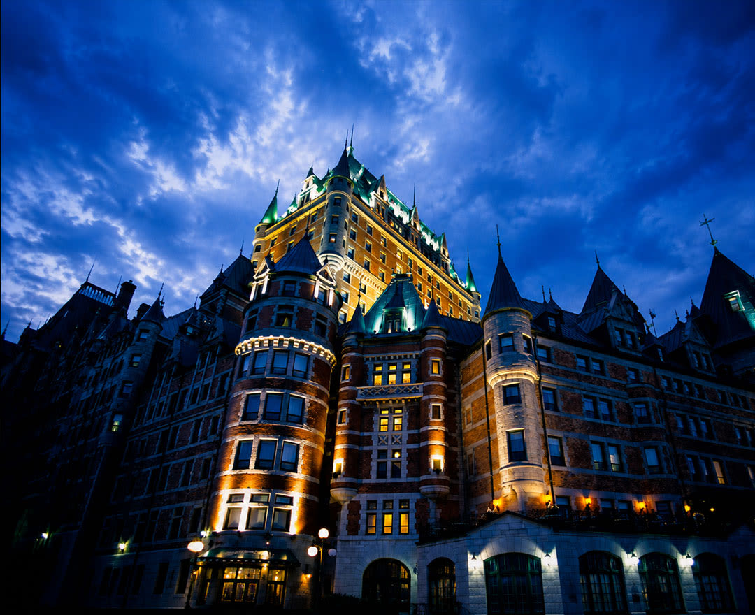 Une vue spectaculaire en contre-plongée du Château Frontenac illuminé à la tombée de la nuit, sous un ciel bleu profond et nuageux