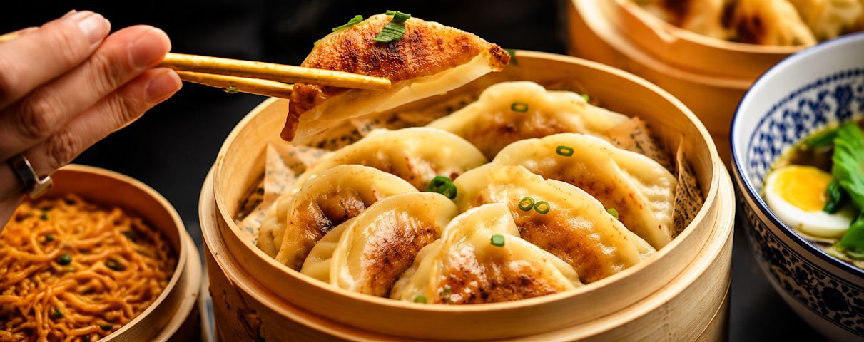 Main de personne tenant un dumpling doré avec des baguettes au-dessus d'un panier vapeur rempli de dumplings, bol de nouilles à côté.
