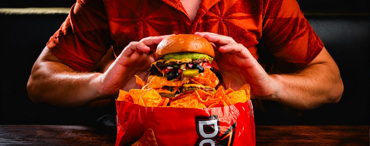 Personne tenant un gros hamburger avec des nachos, portant une chemise rouge. Sac de chips rouge en dessous.