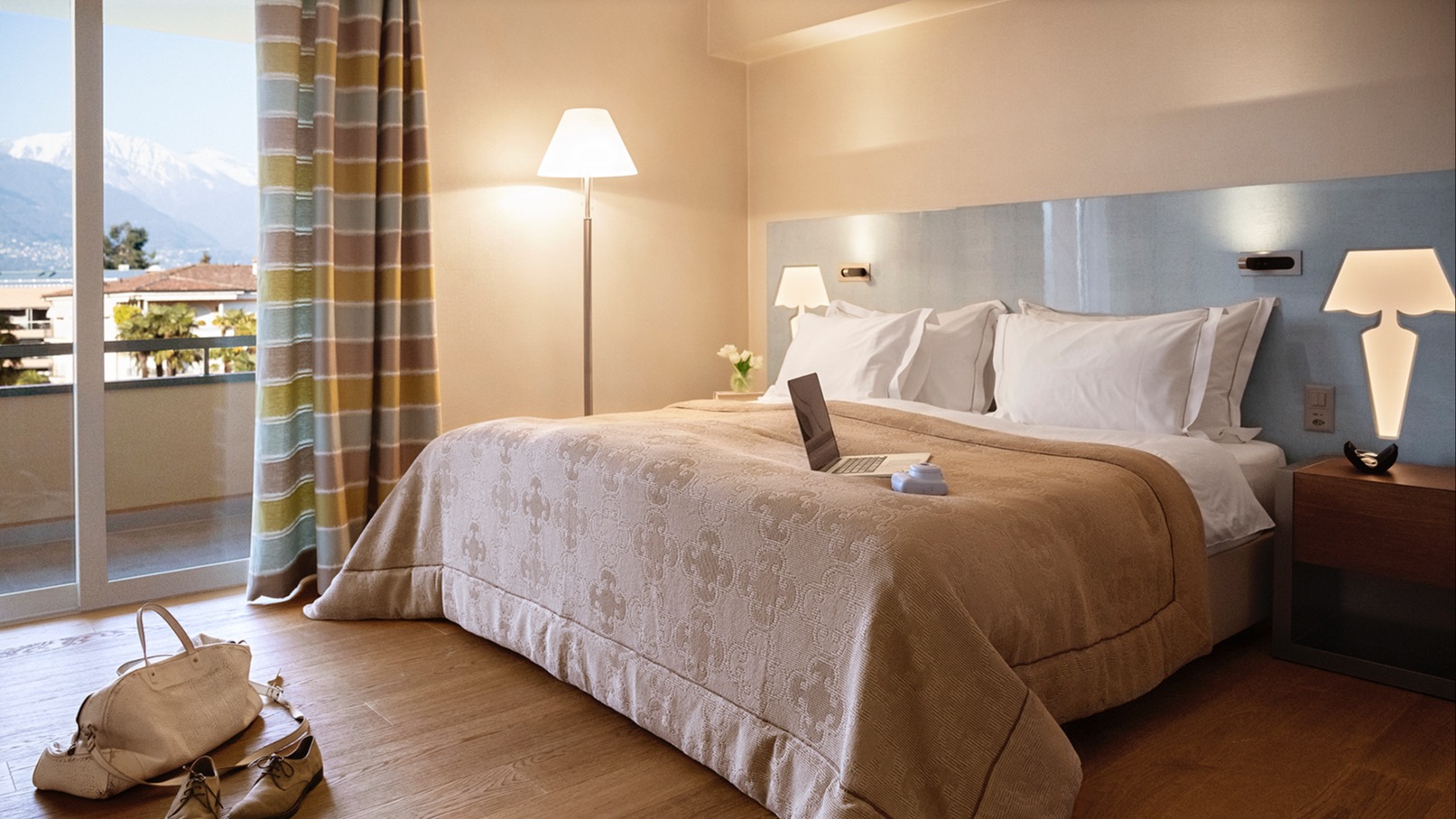 Modern bedroom with large bed, hardwood flooring, striped curtains, balcony with mountain and lake view, and laptop placed on the bed.