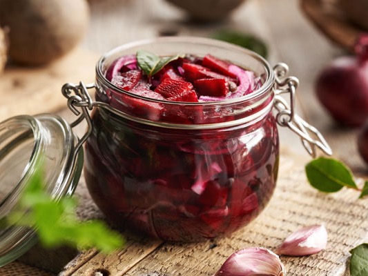 A jar of pickled red vegetables sits on a wooden table, surrounded by fresh ingredients such as onions and herbs.