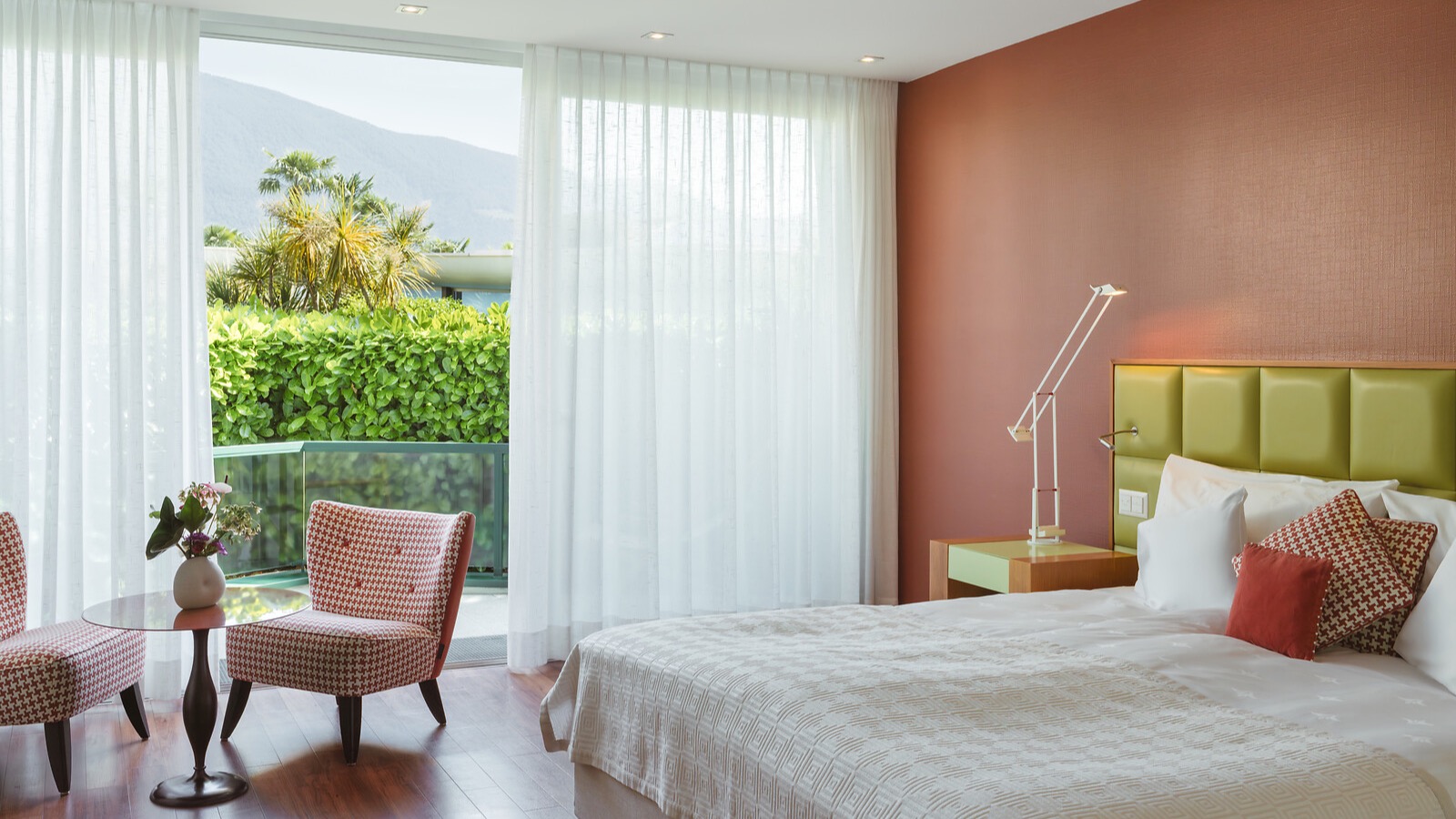 Modernes Hotelzimmer mit terrakottafarbener Wand, grünem Kopfteil, weissen Vorhängen und Blick auf Palmen.