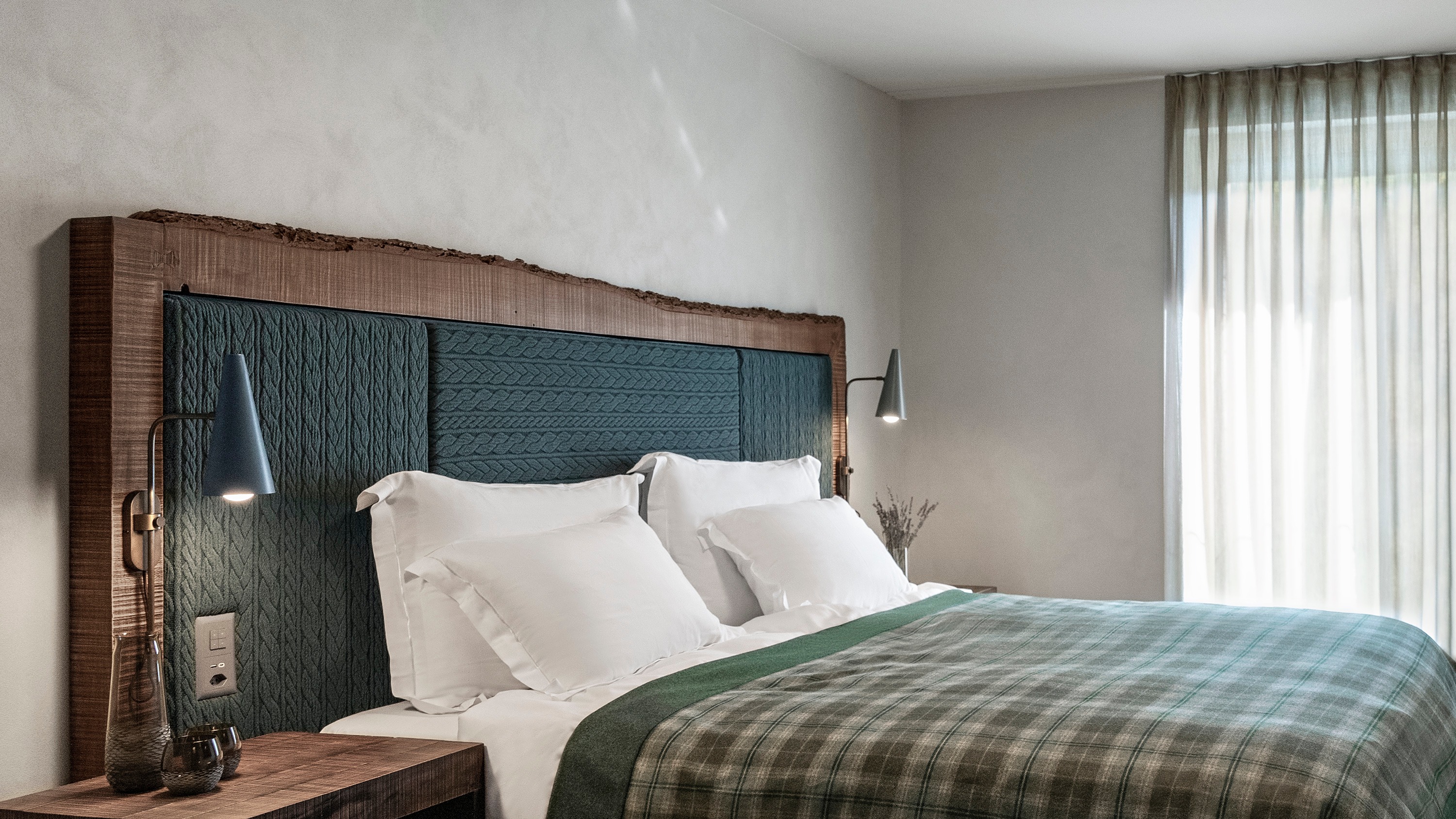 Modern bedroom with teal textured headboard, white bedding, green plaid throw blanket, and wall-mounted reading lamps.
