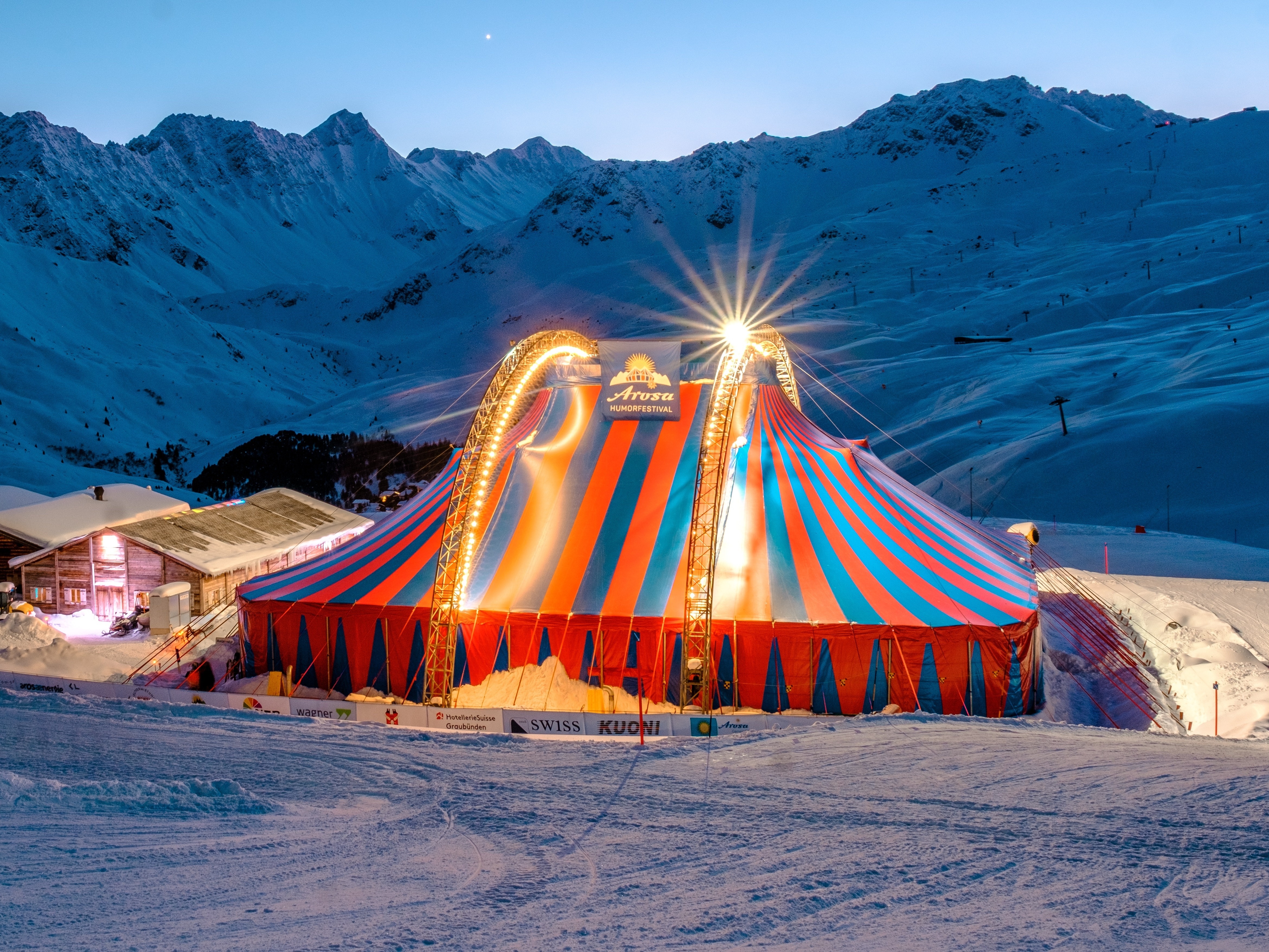 Beleuchtetes Zirkuszelt mit rot-blau gestreiftem Dach vor schneebedeckten Berggipfeln in der Abenddämmerung in einem Skigebiet.