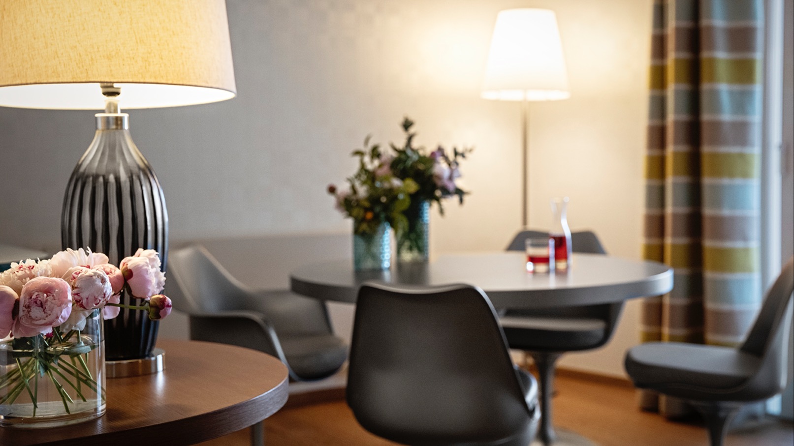 Close-up of a modern dining area featuring a round grey table, four designer chairs, two vases with fresh flowers, striped-base table lamp, pink peonies, and pastel plaid curtains.