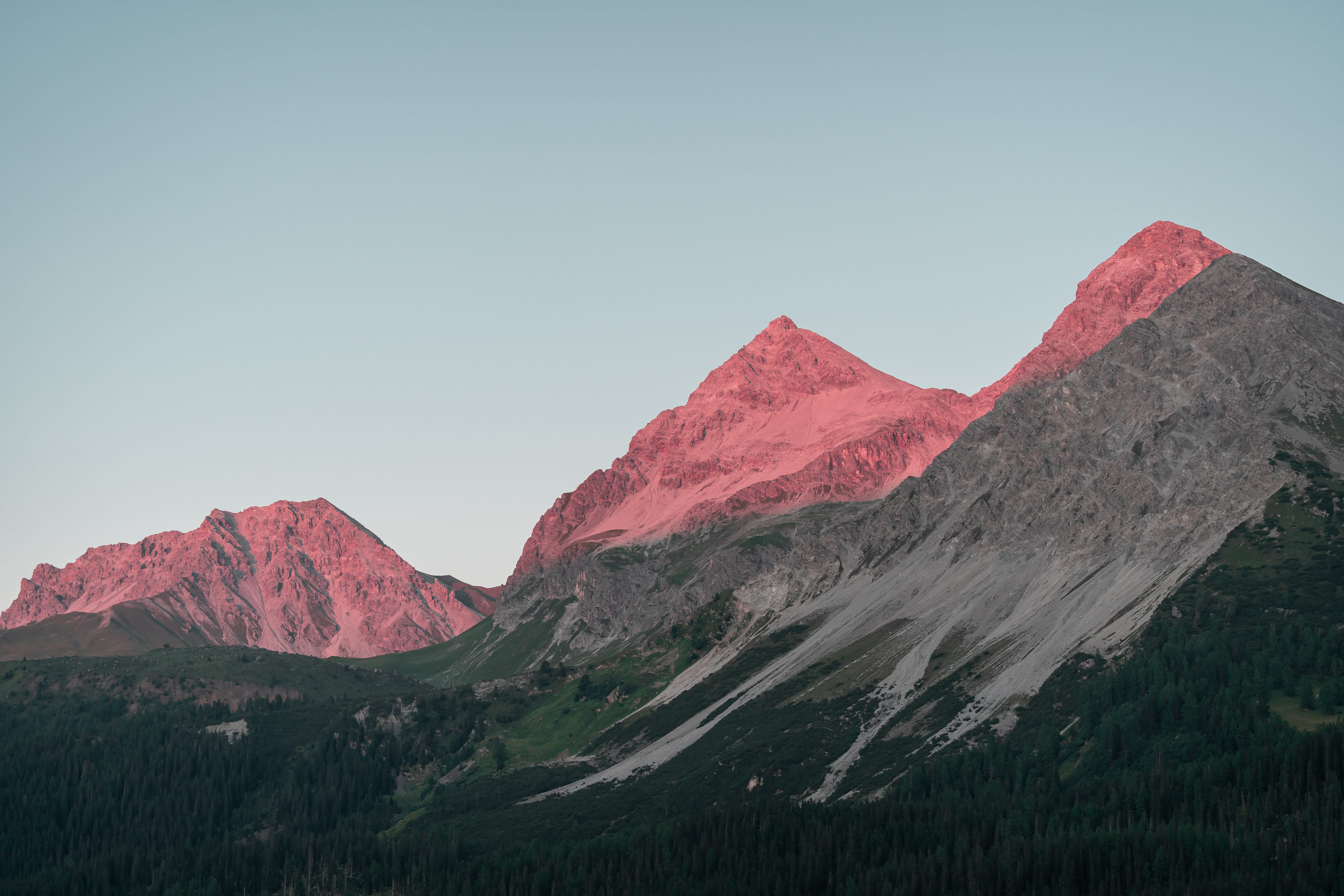 Mountain peaks glowing pink from sunrise or sunset light against a pale blue sky, with gray rocky slopes and green forest below.
