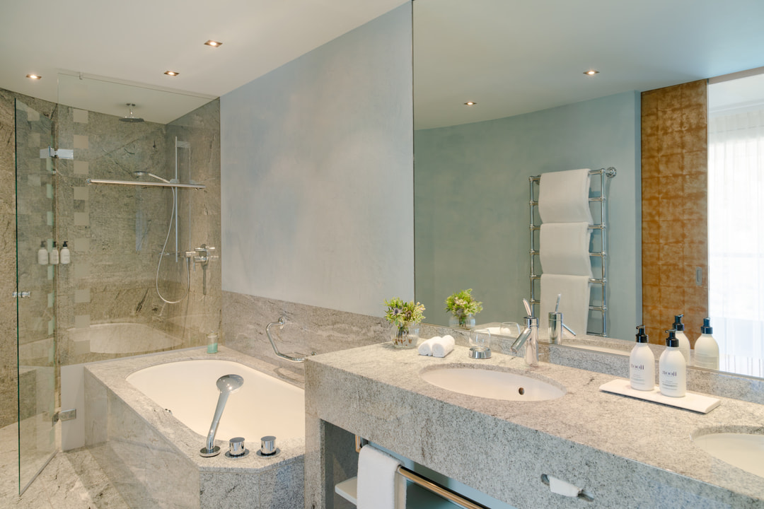 Elegantes Badezimmer mit Granitwaschtisch und Waschbecken, grossem Spiegel, eingebauter Badewanne und verglaster, ebenerdiger Dusche, ergänzt durch einen Handtuchwärmer und hellblaue Wände.