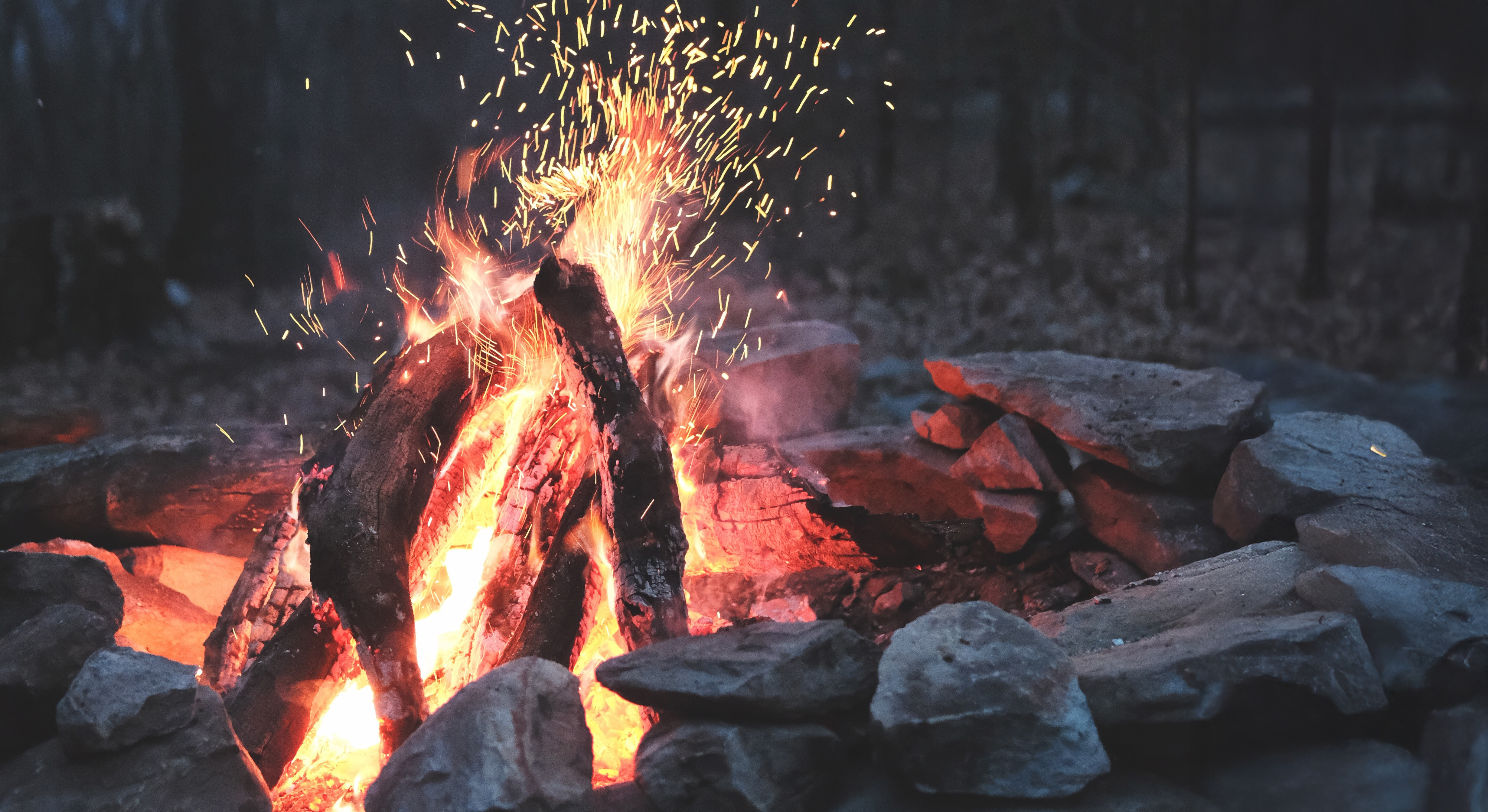 Campfire with burning logs and bright orange flames shooting sparks upward, surrounded by a stone ring against a dark background.