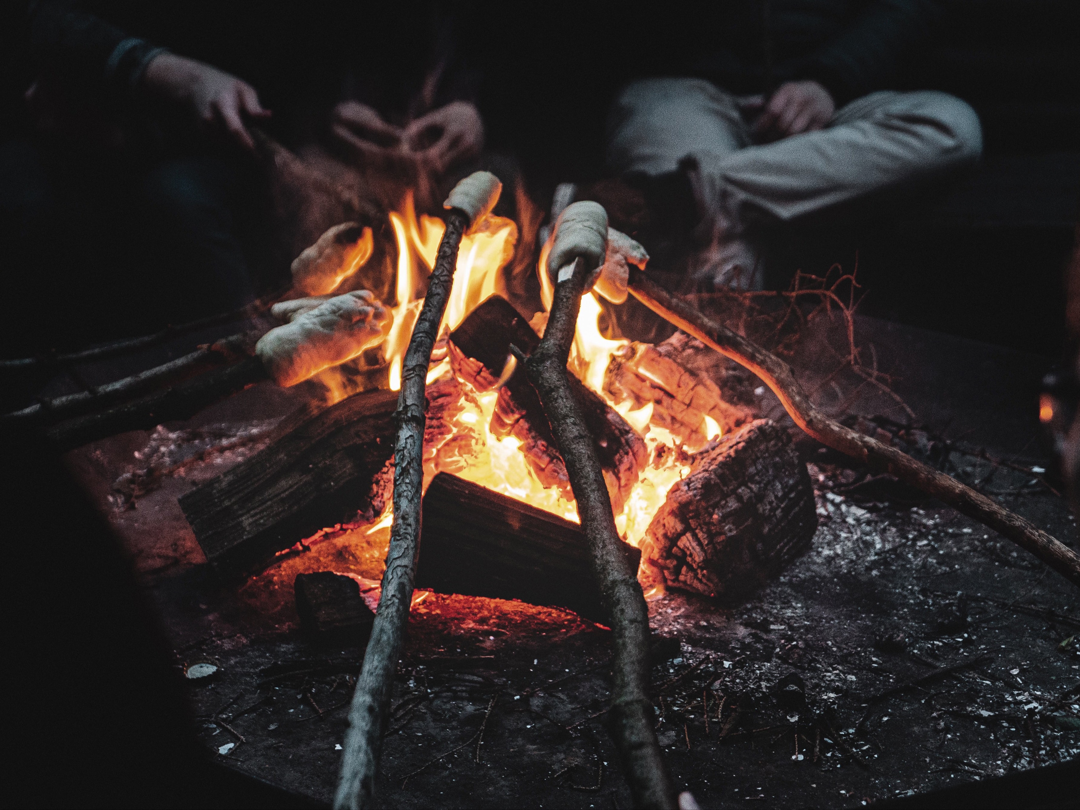 People warming hands around a glowing campfire with orange flames and burning logs at night.