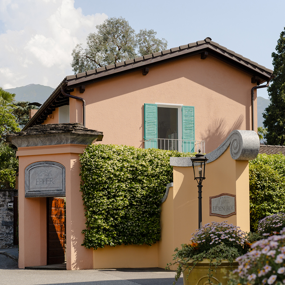 Pfirsichfarbenes Haus mit türkisfarbenen Fensterläden, umgeben von Palmen und Blumen, mit Steintor und Laterne.