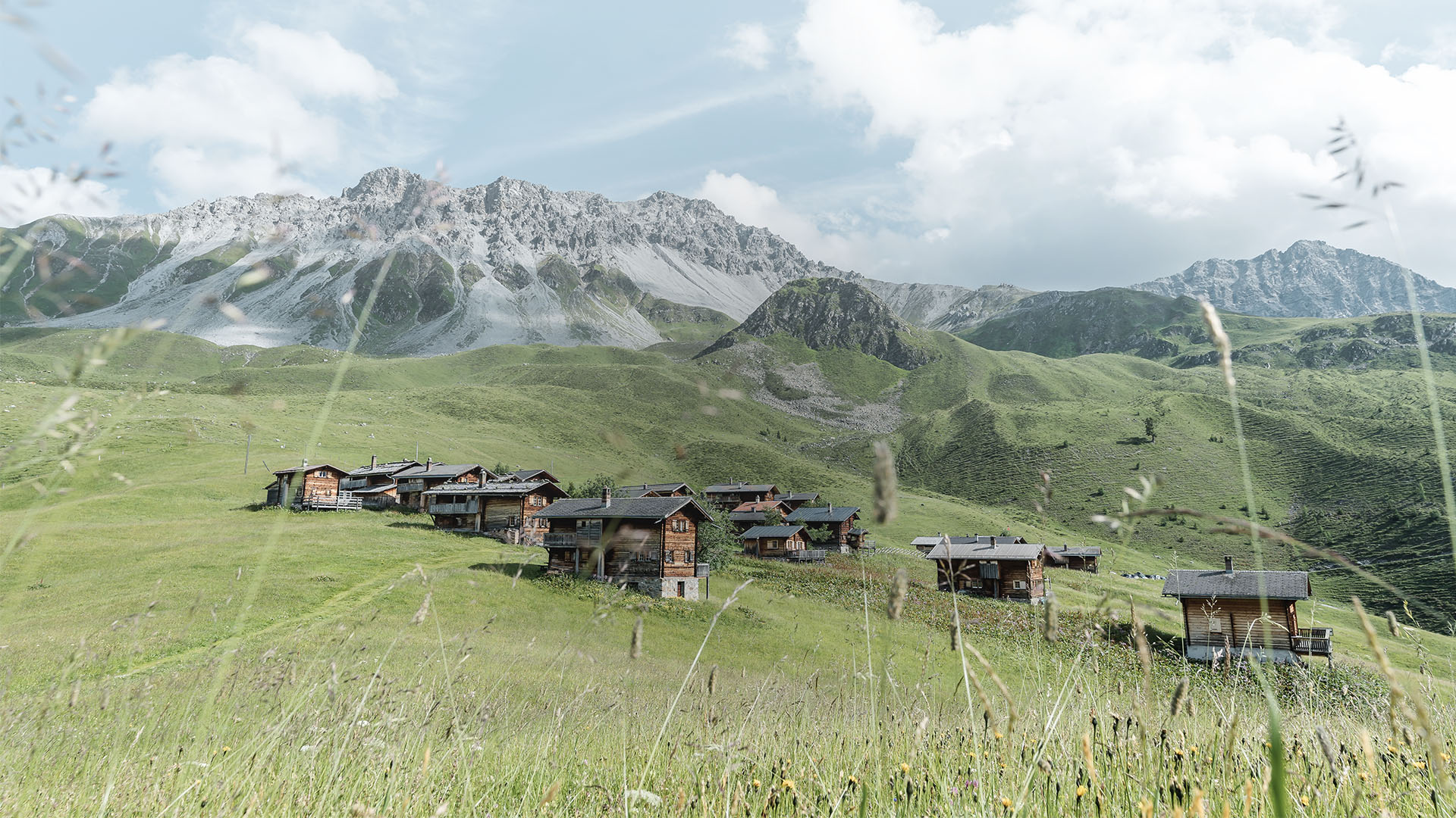 Ein Alpendorf mit Holzchalets, die eingebettet in grüne Wiesen unter schneebedeckten Berggipfeln und einem bewölkten blauen Himmel liegen.