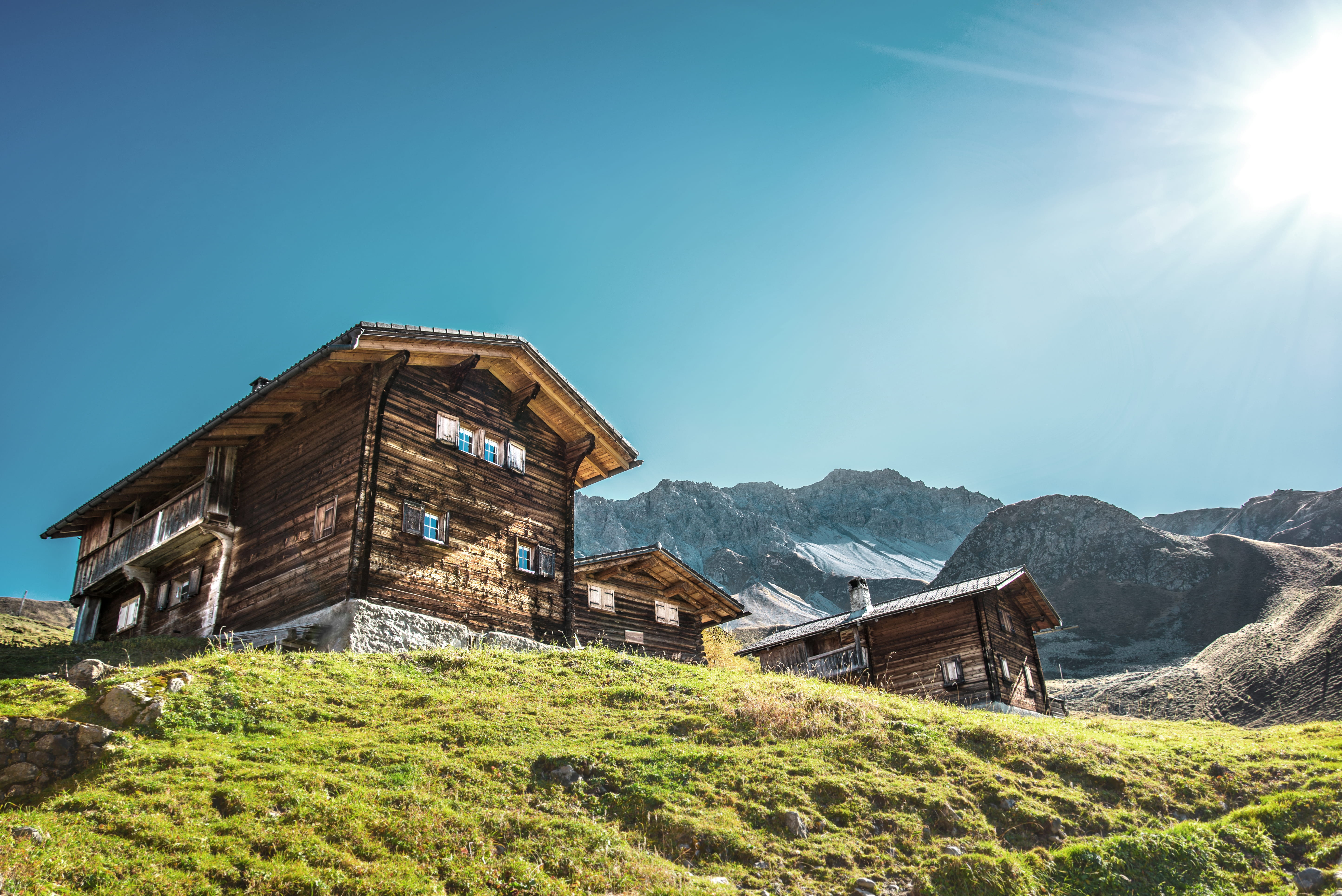 Traditional wooden alpine chalets on a green hillside with snow-capped mountains and blue sky in the background.