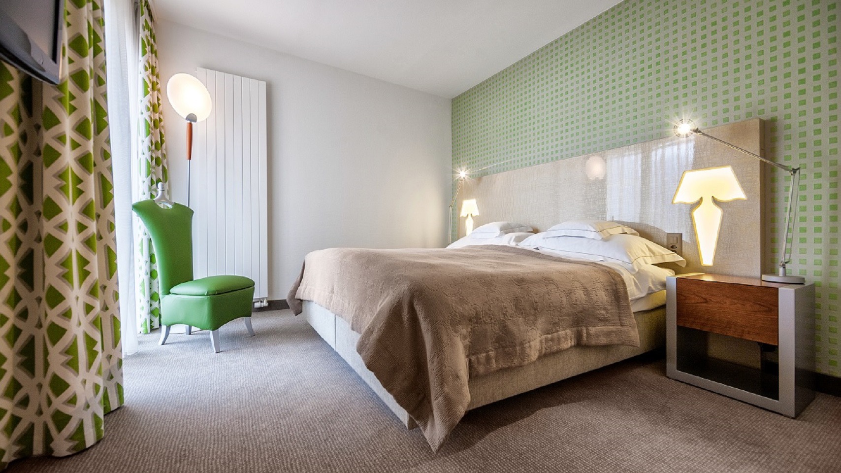 Modern bedroom featuring a green patterned accent wall, beige bedspread, double bed with white linens, designer lamps, and a green accent chair.