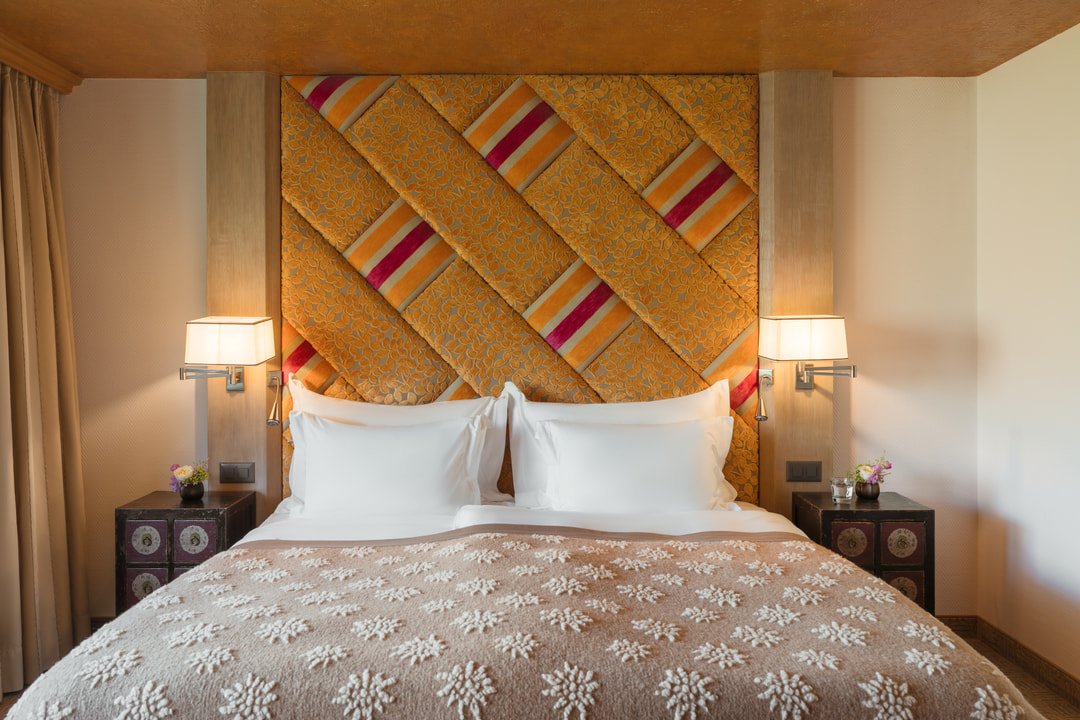 Elegantes Hotelzimmer mit grossem Doppelbett, markantem orangefarbenem Kopfteil mit Muster, weissen Kissen und beiger Tagesdecke mit Blumenmotiv, ergänzt durch Nachttische und warme Wandleuchten.