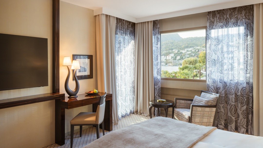 Elegant hotel room with desk, modern lamp, TV, sitting area, and large windows overlooking the lake and green hills.