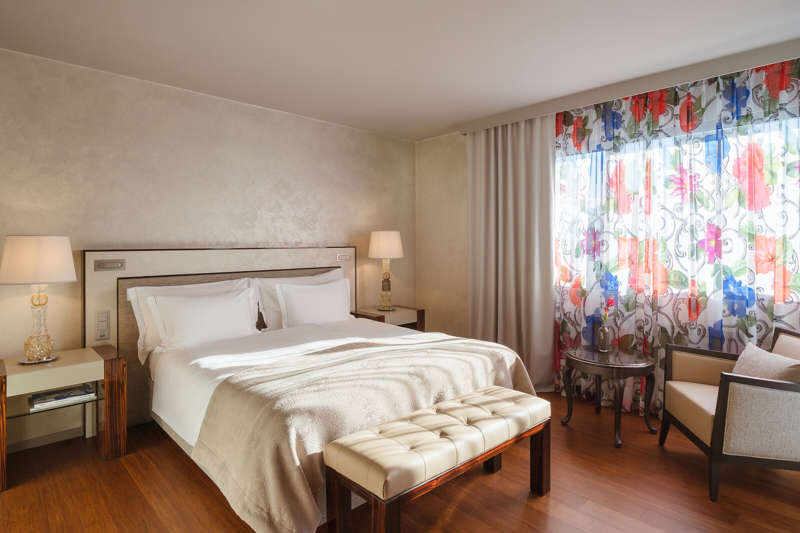 Großzügiges Doppelzimmer mit gemütlichem Sessel, Beistelltisch mit Obst, Tageslicht durch bodentiefe Fenster und dezentem Design.