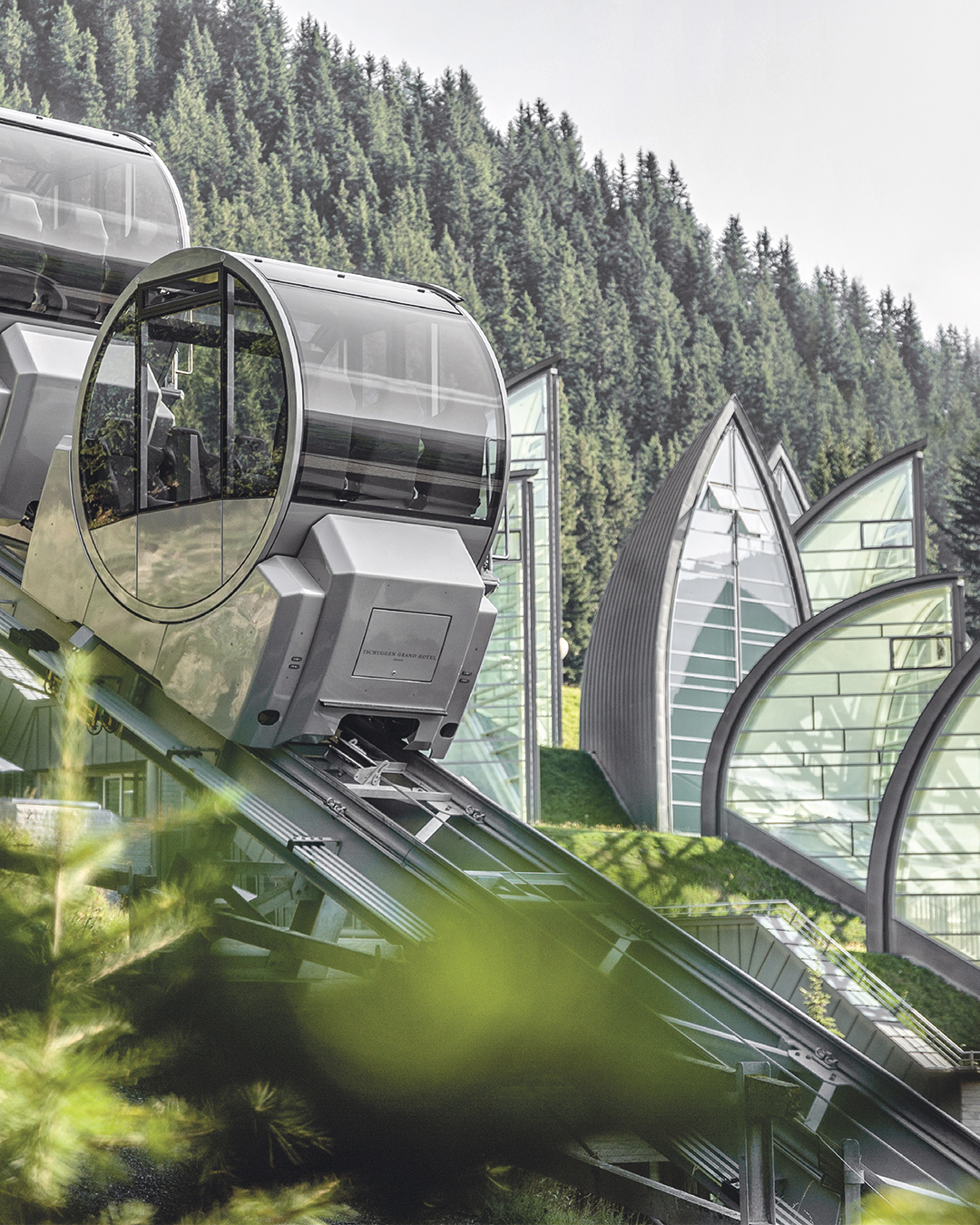 Moderne Seilbahngondeln mit Glasfenstern fahren über eine Bergstation mit grünen Dächern vor einem dichten Nadelwald.