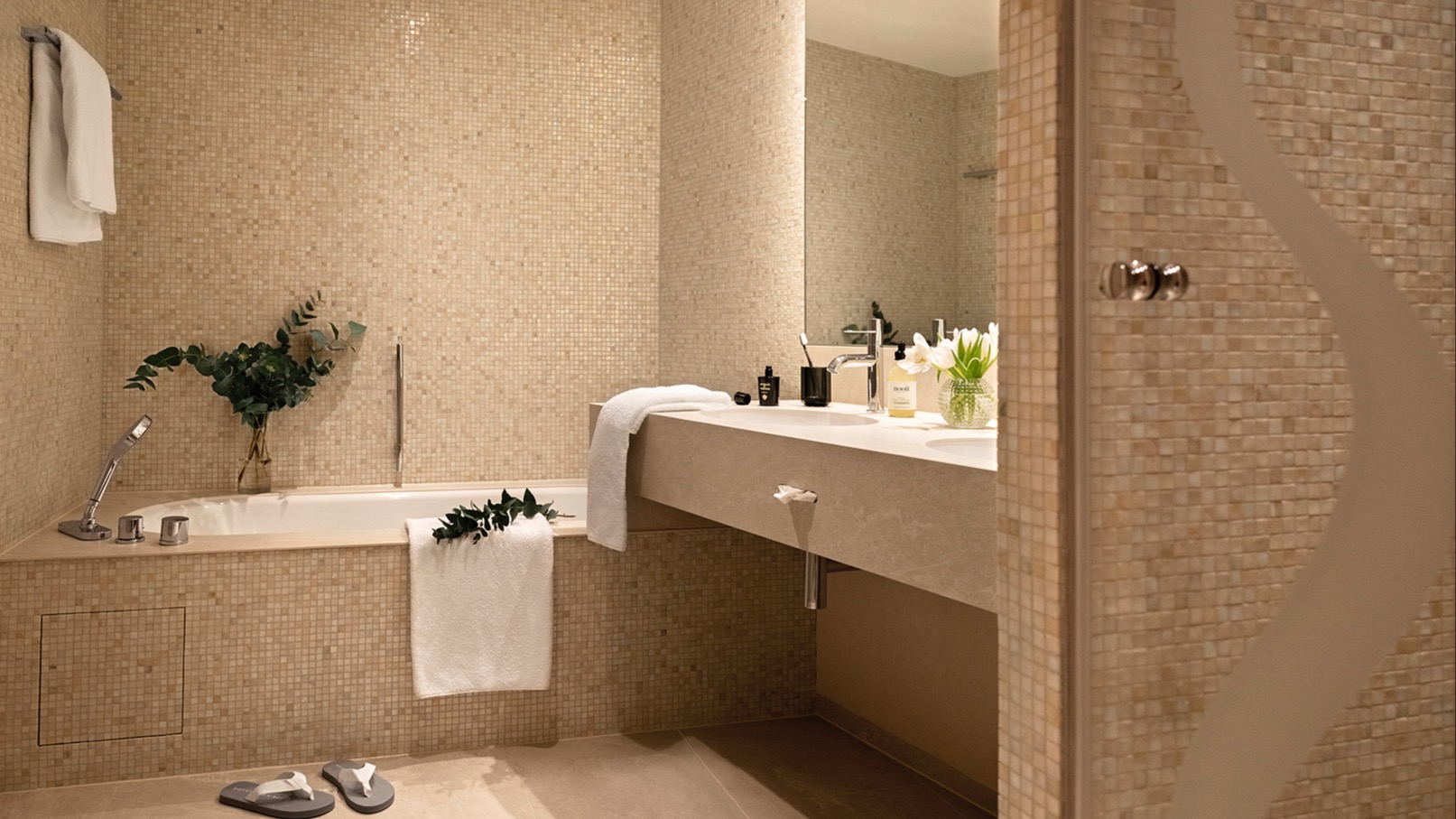 Modern bathroom with beige mosaic tiles, built-in bathtub, double sink vanity, large mirror, and decorative touches including fresh eucalyptus branches and white towels.
