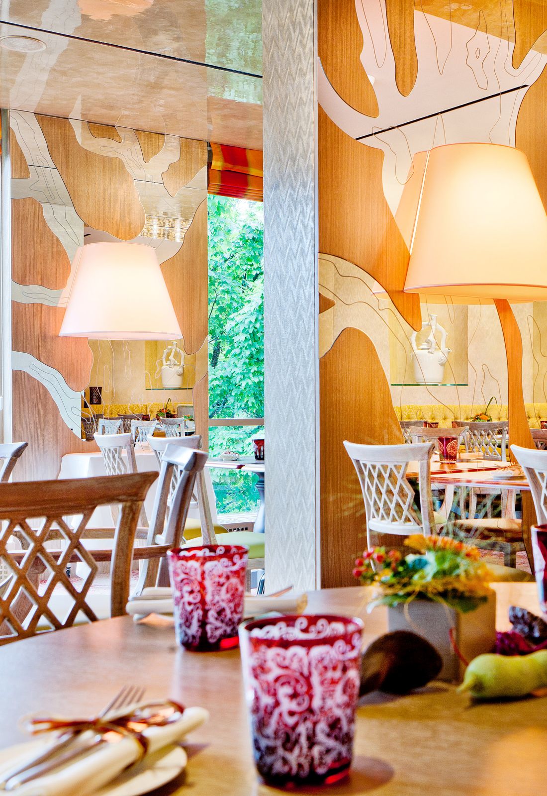Modern restaurant interior with artistic wood panels, white pendant lamps, lattice chairs, and colorful glassware on tables.