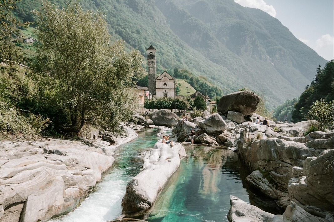 Türkisfarbenes Flussbecken zwischen hellen Felsen im Maggiatal, mit einer Kirche und bewaldeten Bergen im Hintergrund.