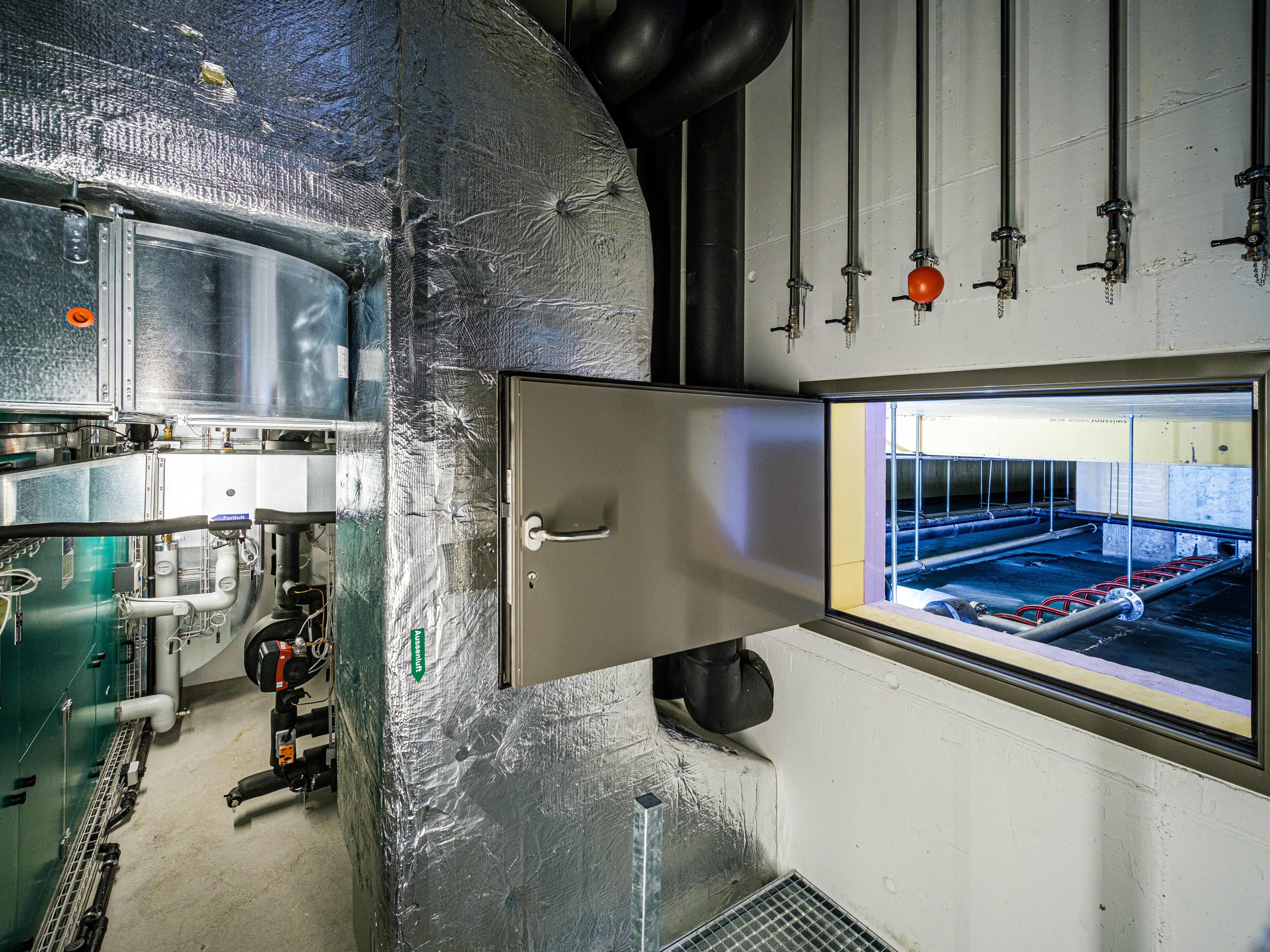 Technikraum des Eisspeichers im Valsana mit Lüftungs- und Rohrleitungssystemen; durch eine geöffnete Wartungsklappe ist der Eisspeicher mit seinen Leitungen sichtbar.
