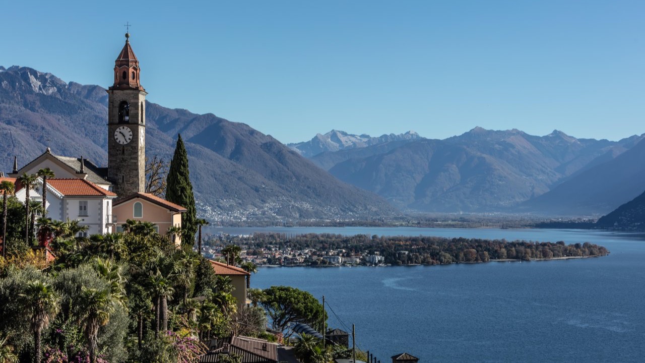 Church with a bell tower overlooking Lake Maggiore with a view of the Brissago Islands and the surrounding mountains.