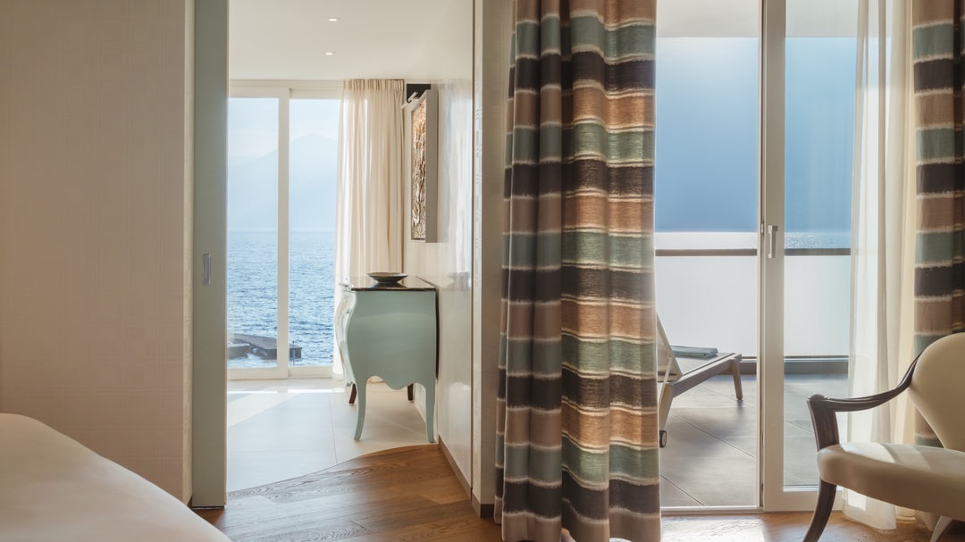 Helles Schlafzimmer mit Zugang zum Balkon, bodentiefen Fenstern, seitlichem Meerblick, gemusterten Vorhängen und stilvollem Mobiliar in Pastelltönen.