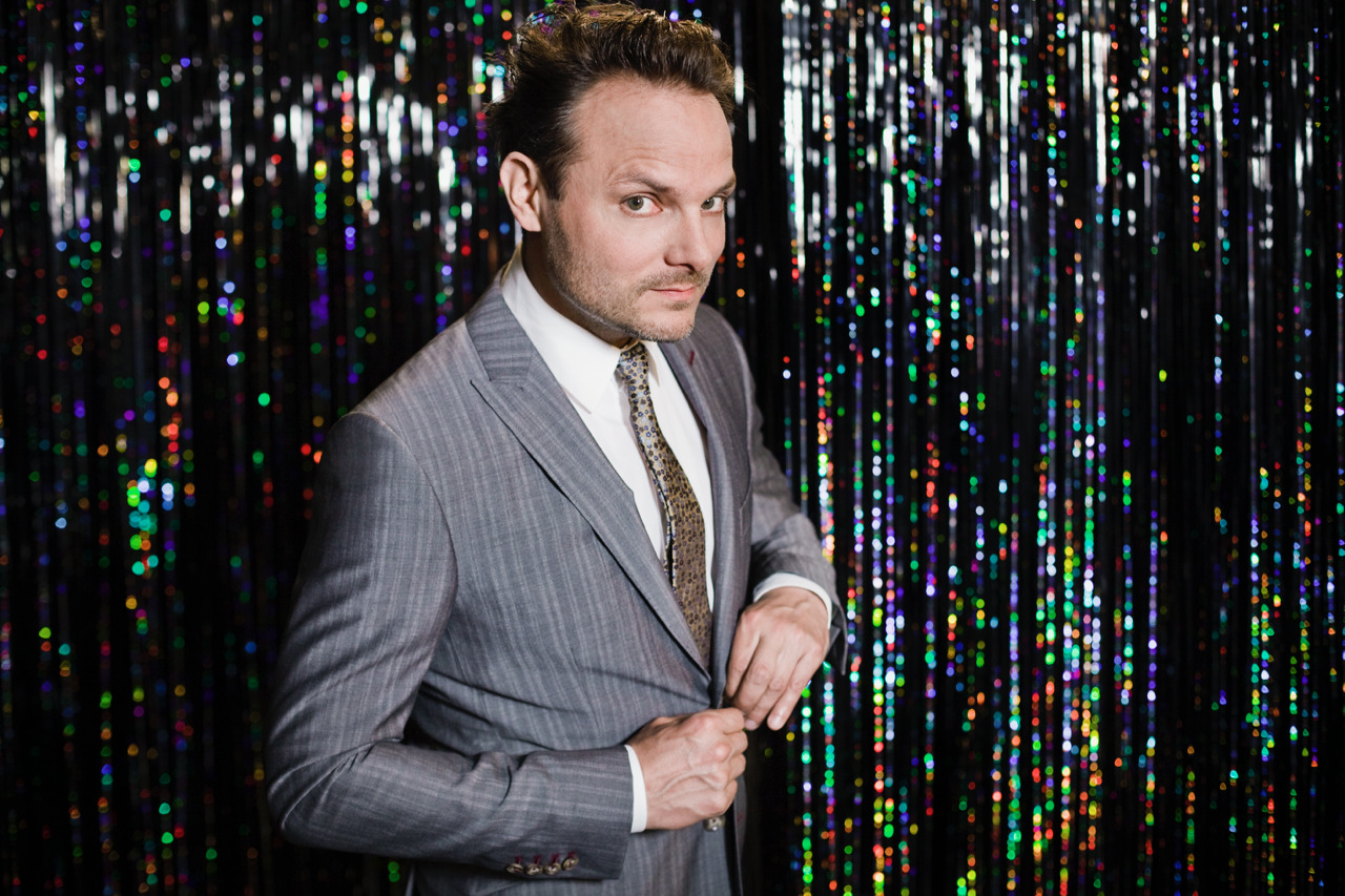 Dominic Deville in a grey suit in front of a shimmering, colourful tinsel curtain.