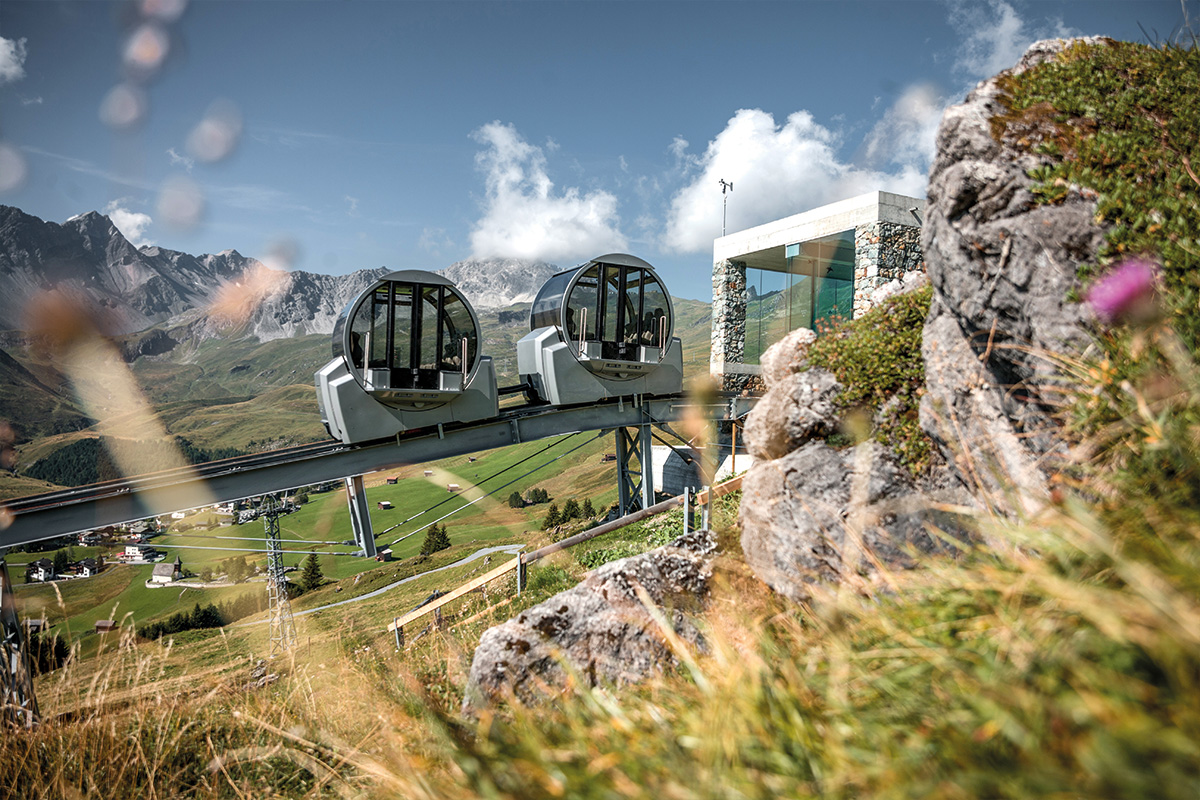 Moderne Seilbahnstation mit runden Glasfenstern in alpiner Berglandschaft unter blauem Himmel mit weissen Wolken.