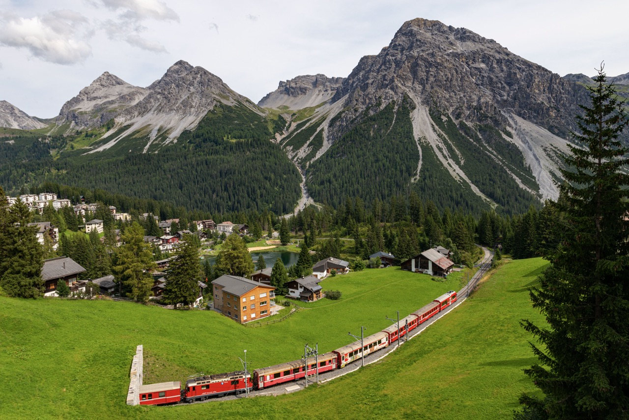 Red train traveling through green alpine valley with dramatic mountain peaks, forests, and village buildings in background.