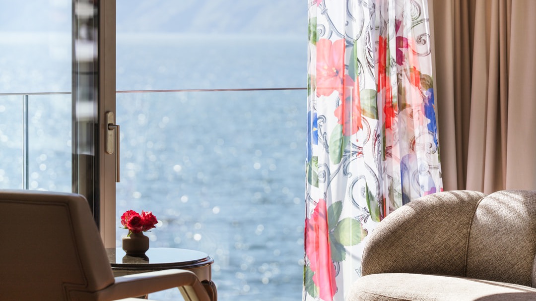 Detailaufnahme eines Sitzbereichs mit floralen Vorhängen, kleinem Beistelltisch mit Vase und Blick auf das glitzernde Wasser des Sees.