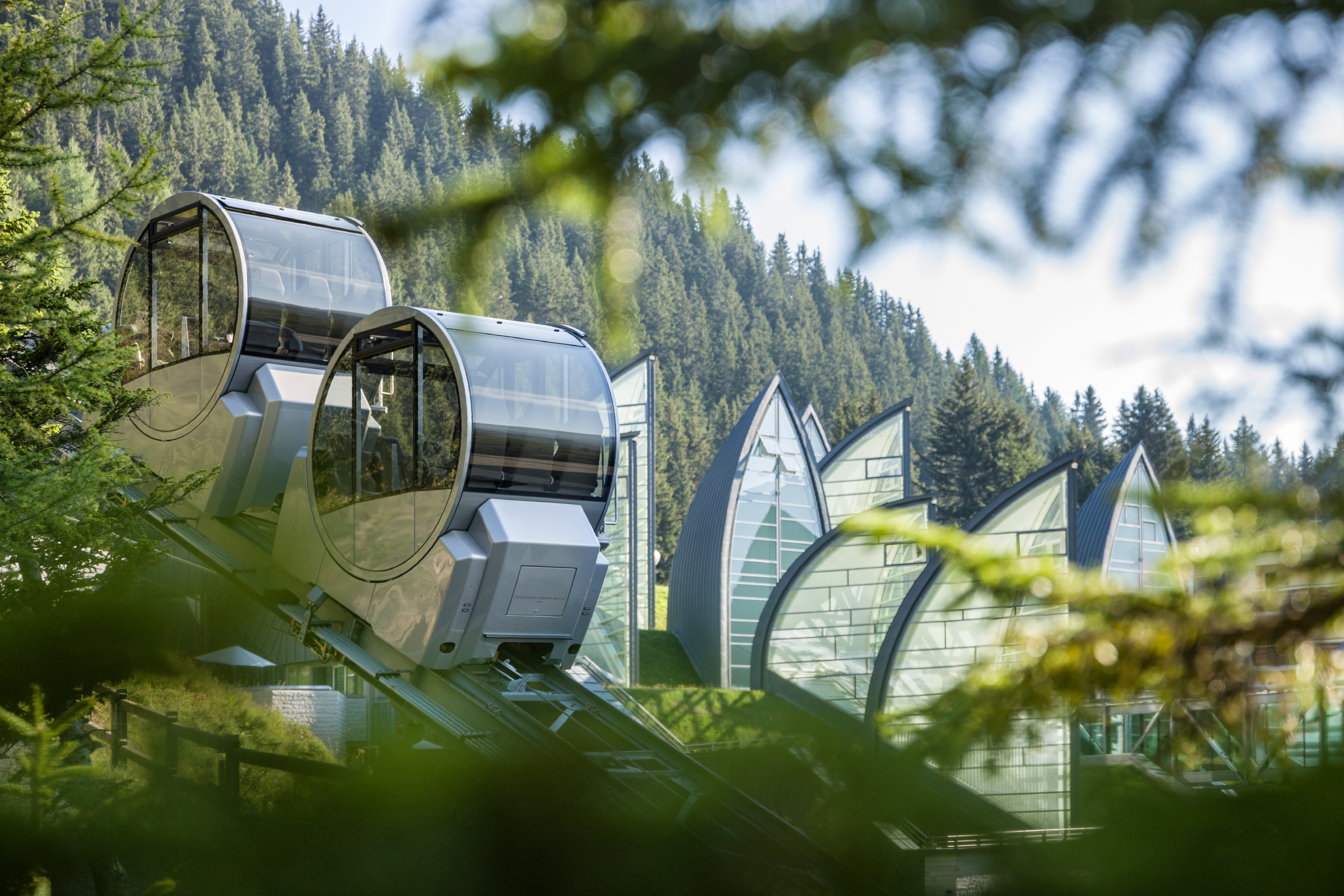 Moderne Gondelbahn-Kabinen mit Glasfenstern, die über weiße Gebäude in A-Form  hinaufschweben, umgeben von einem immergrünen Wald.